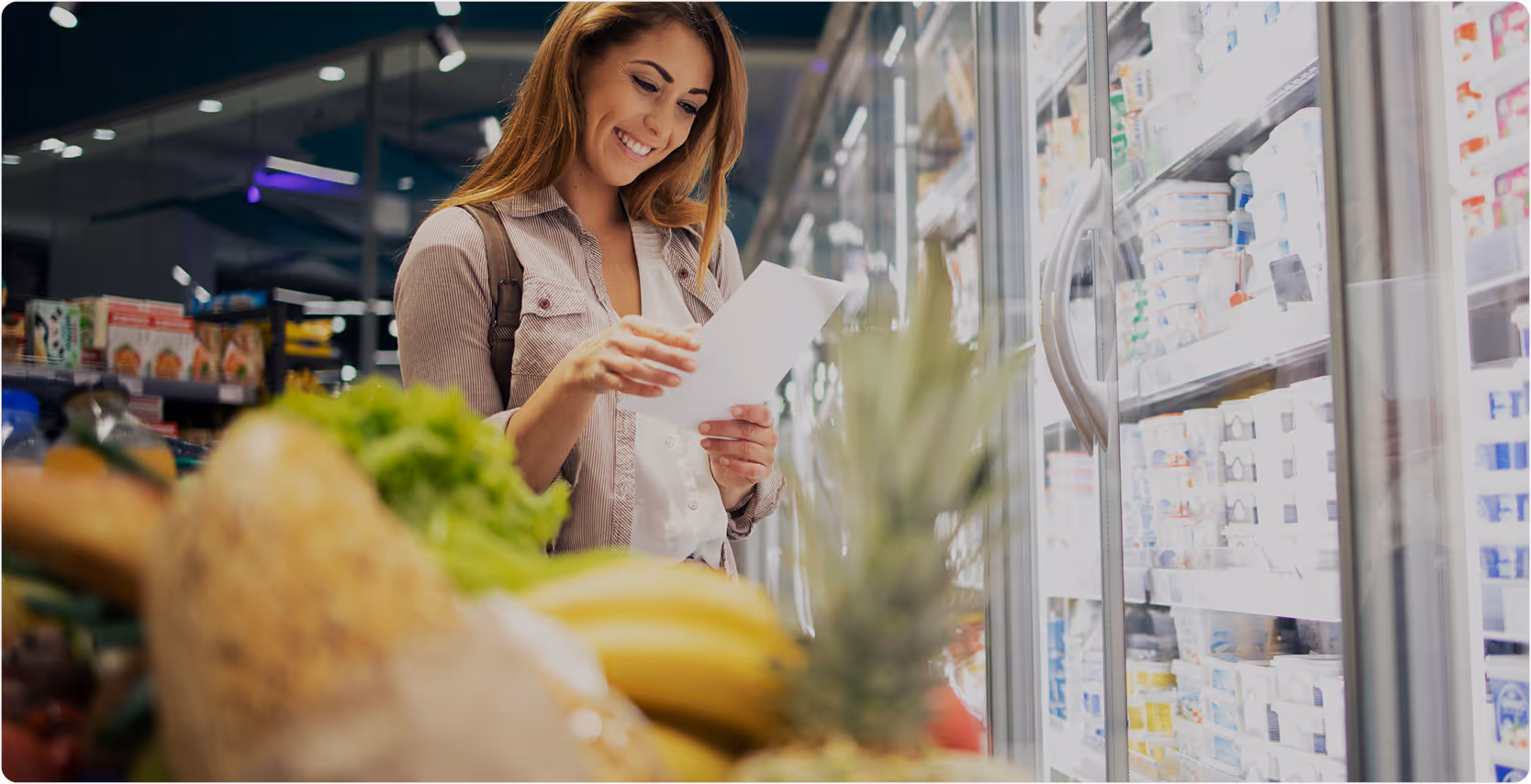 Woman smiling while reading a shopping list in front of refrigerated grocery shelves.