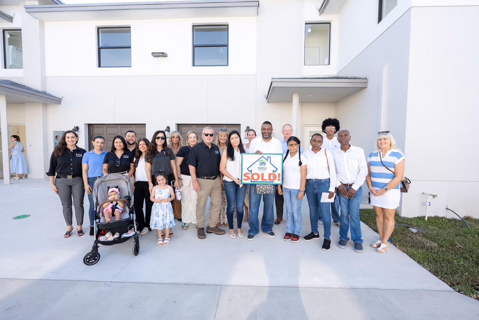 Families and BBI associates gathered outside a newly completed BBI Village home, holding a Habitat for Humanity sold sign