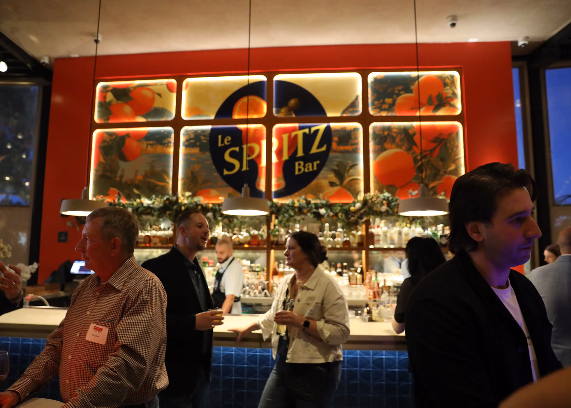 Interior of Le Spritz Bar with people socializing and drinking near the bar counter, illuminated by hanging lights and a large sign with oranges and the bar's name.