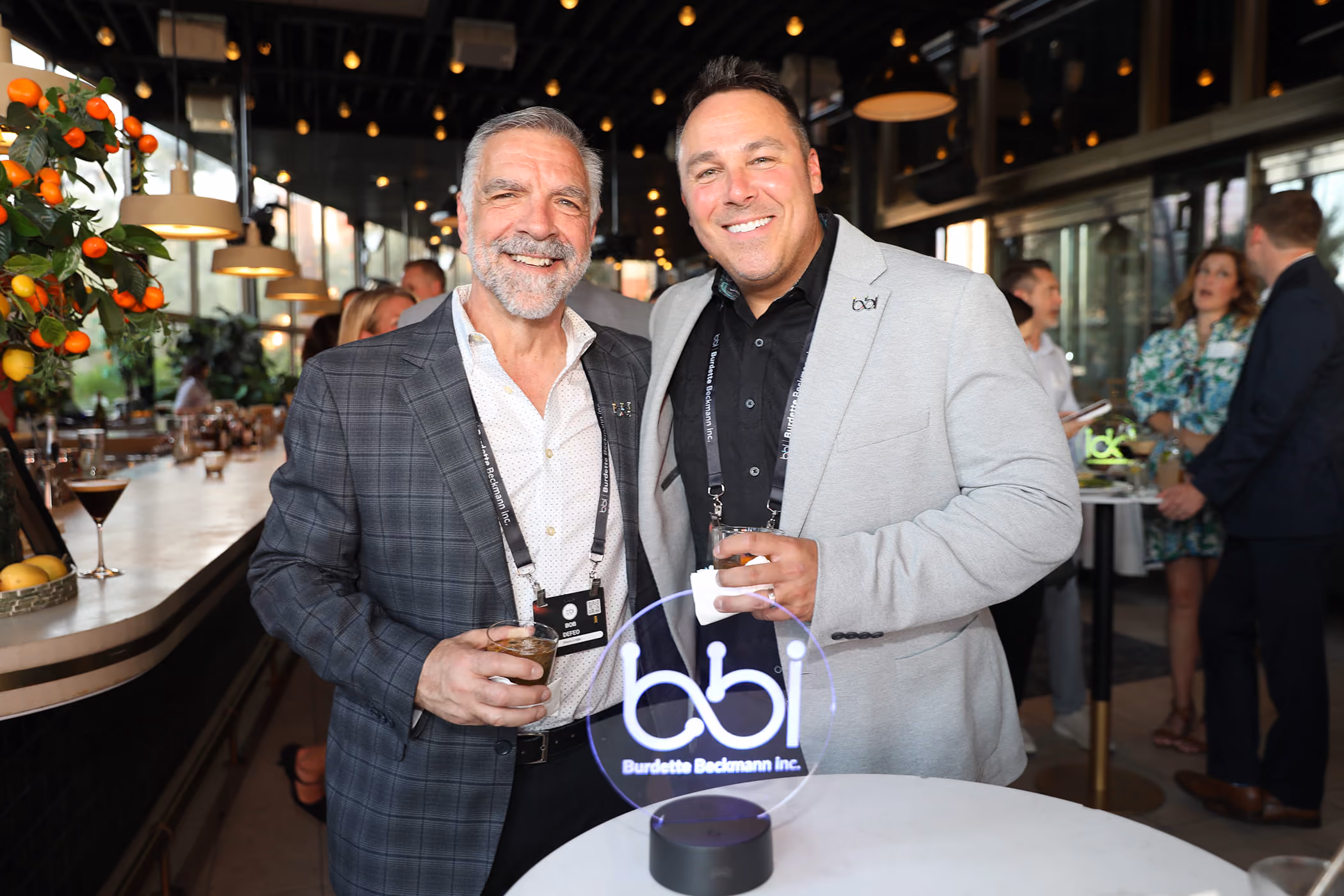 Two men smiling and posing for a photo at an indoor event, each holding a drink, with a Burdette Beckmann Inc. light-up sign on the table in front of them.