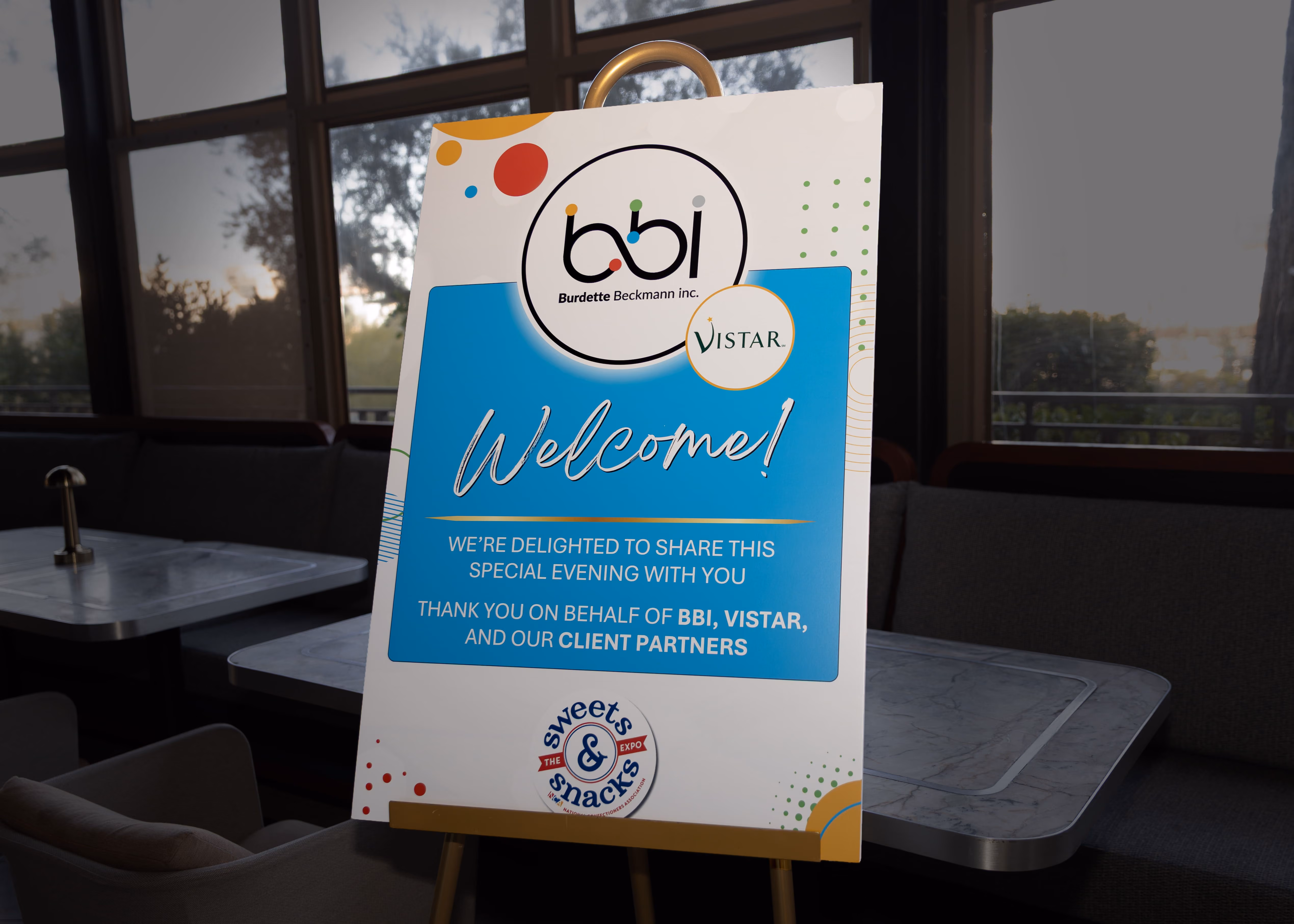 Welcome sign on an easel in a seating area with tables and chairs, displaying logos for BBI, Vistar, and Sweets & Snacks Expo, thanking guests for a special evening.