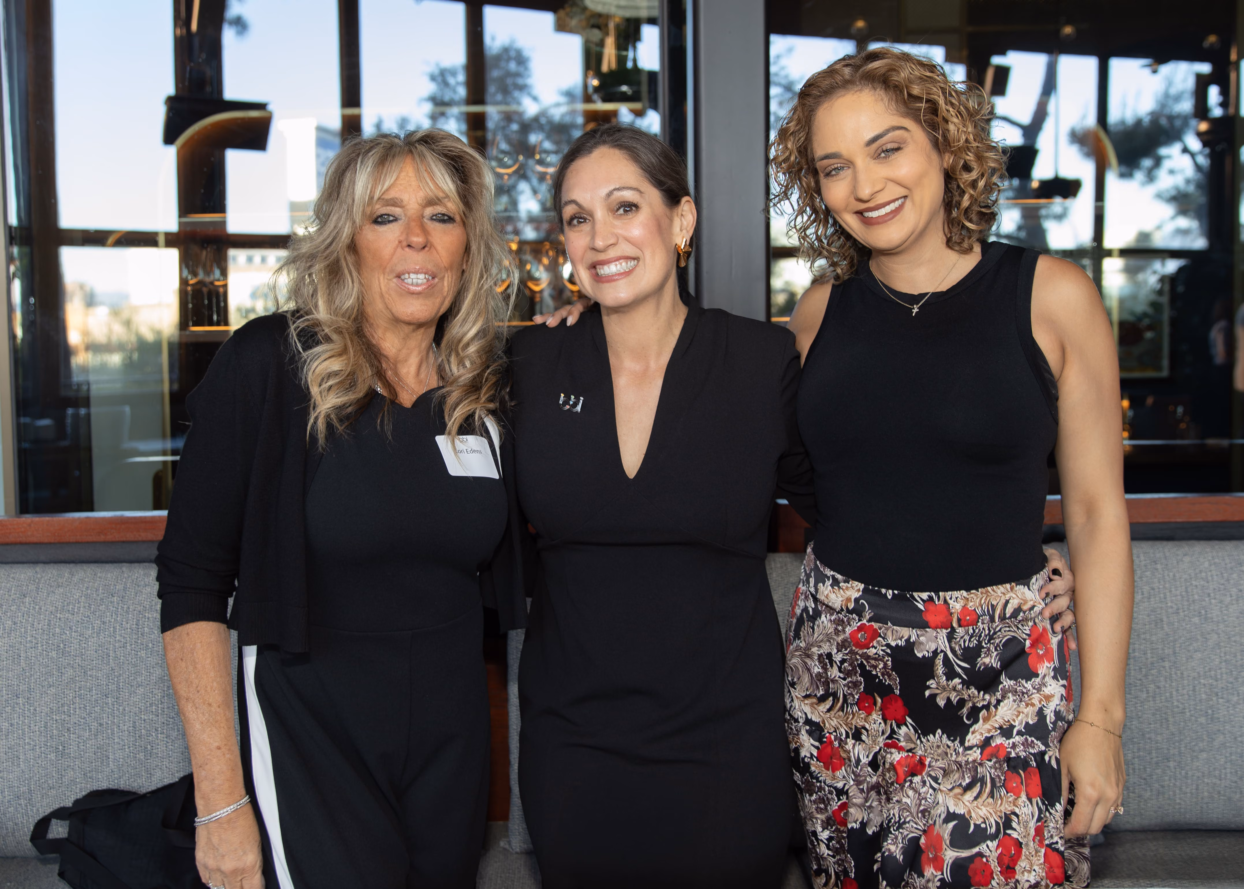 Three women standing close together indoors in front of large windows, smiling at the camera; the woman on the left has long blonde hair and wears black with a cardigan, the woman in the middle has her arm around the other two and wears a black dress, and the woman on the right has curly hair and wears a sleeveless black top with a floral skirt.