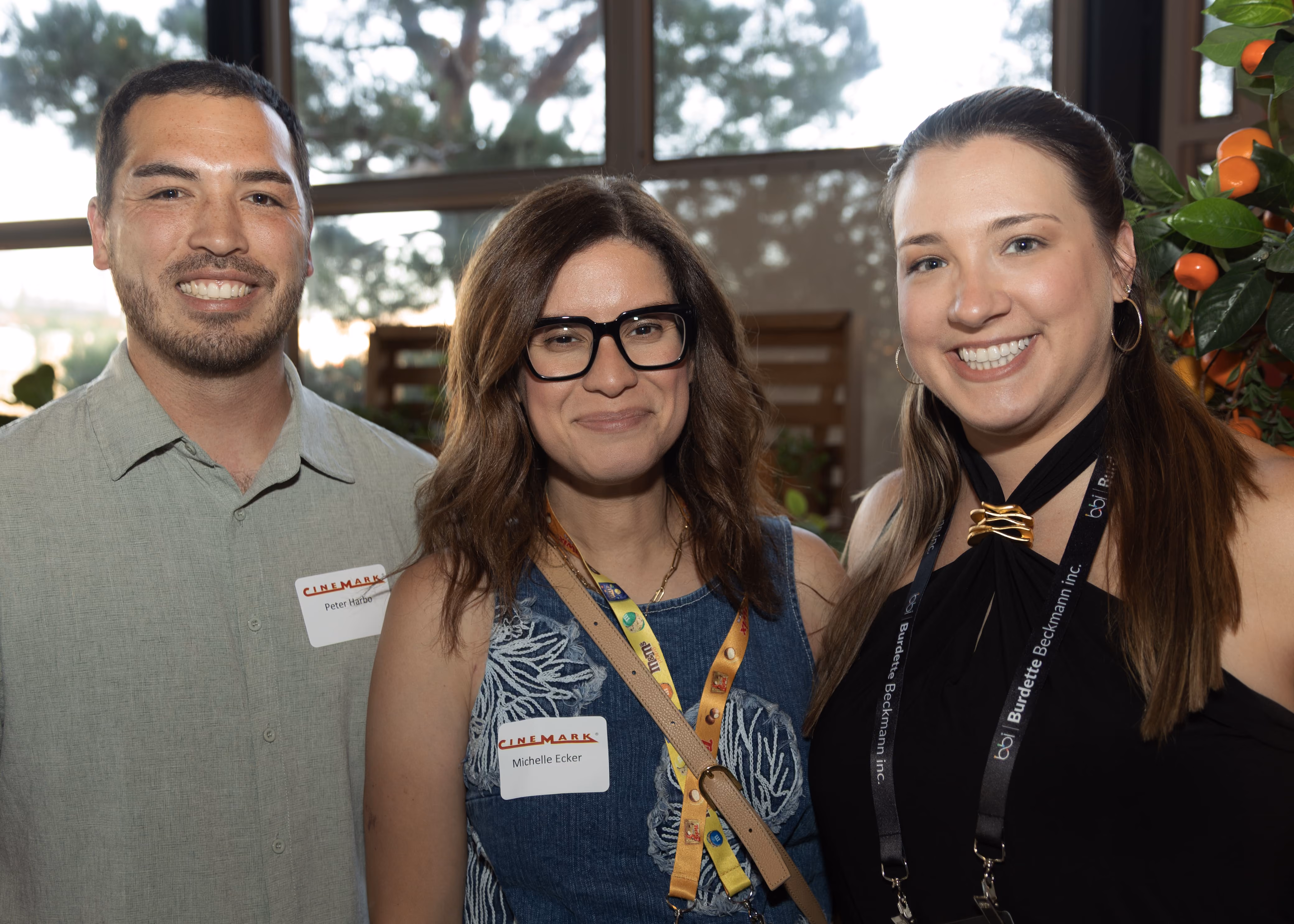 Three smiling people standing closely together indoors near windows; the man on the left wears a light green shirt and a name tag, the woman in the middle has glasses and a blue sleeveless top with a name tag and lanyard, and the woman on the right wears a black sleeveless top with a bow and lanyard.