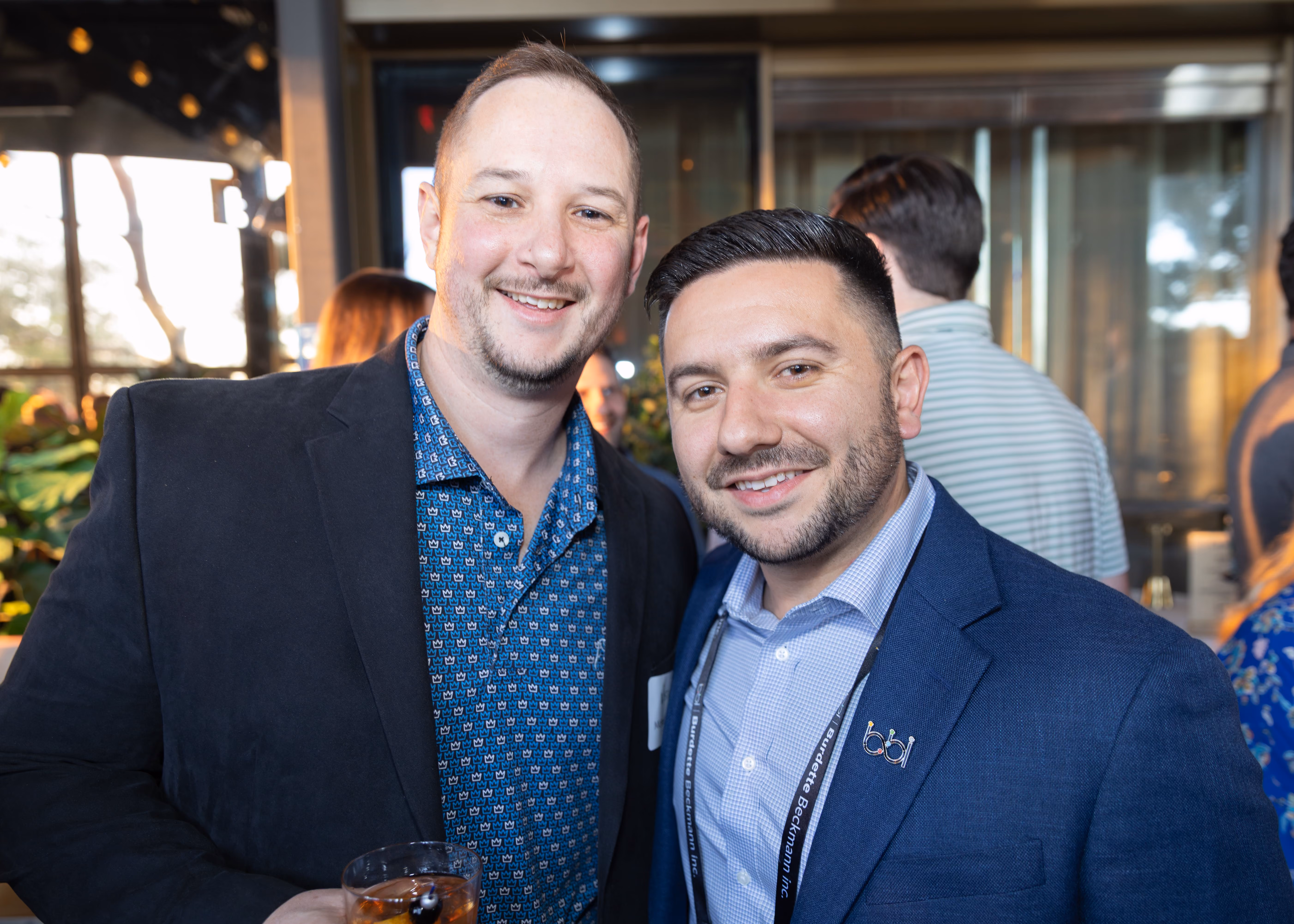 Two men smiling and posing closely together at a social event, one holding a drink, both wearing blue blazers over shirts.