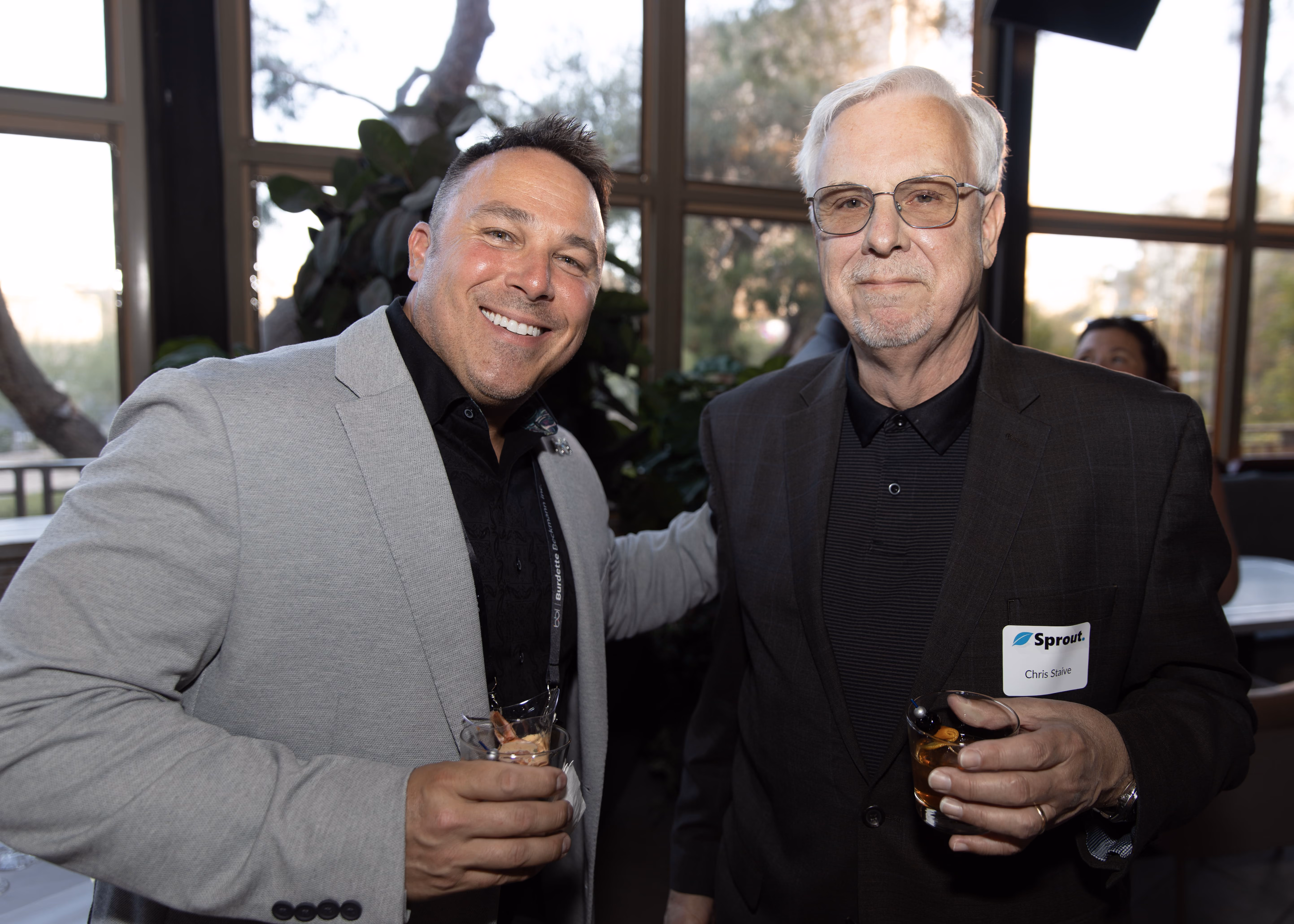 Two men smiling and holding drinks at an indoor social event with large windows in the background.