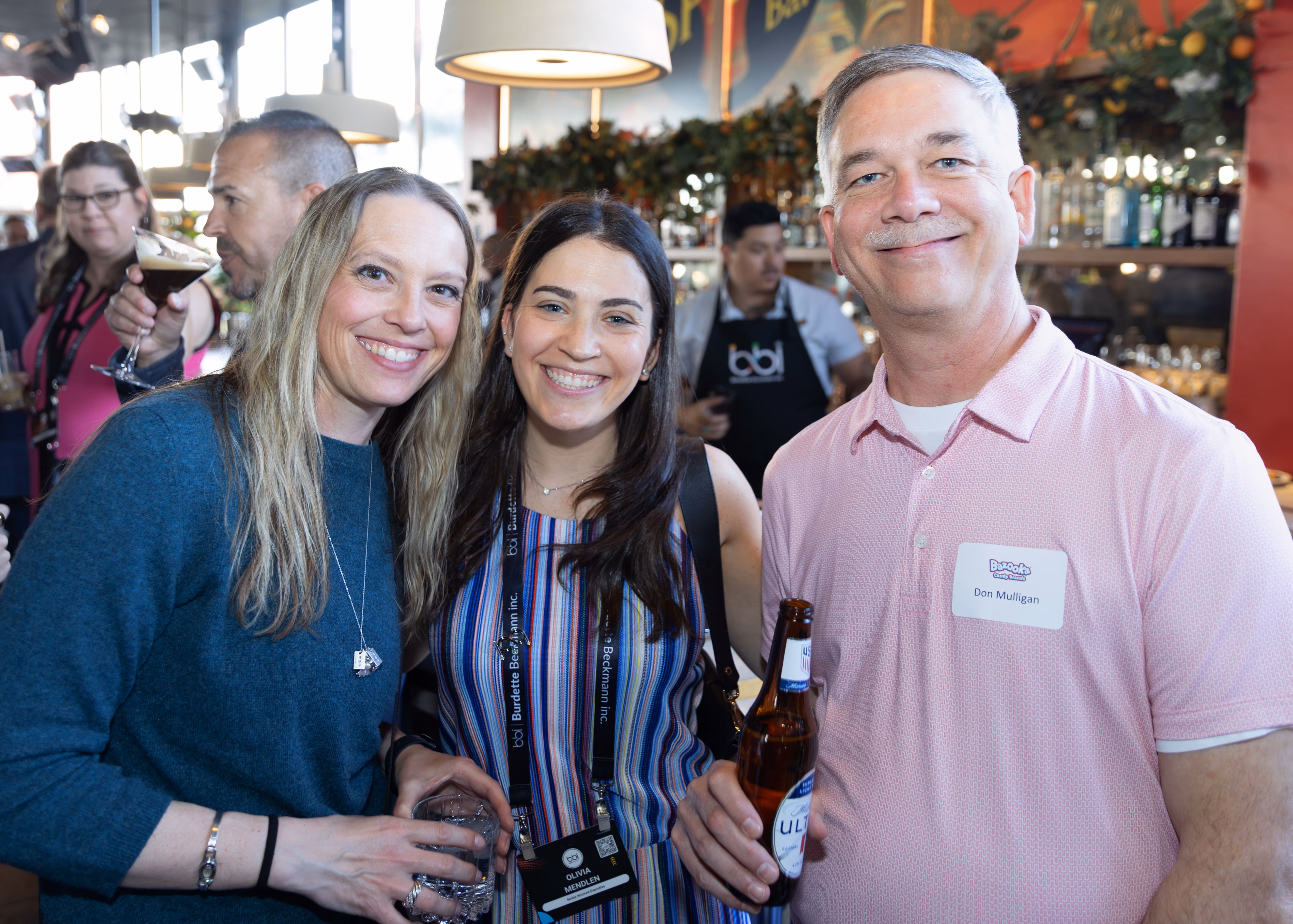 Three people smiling indoors at a social event; two women on the left and a man on the right holding a beer bottle, with a busy background including other guests and a bartender.
