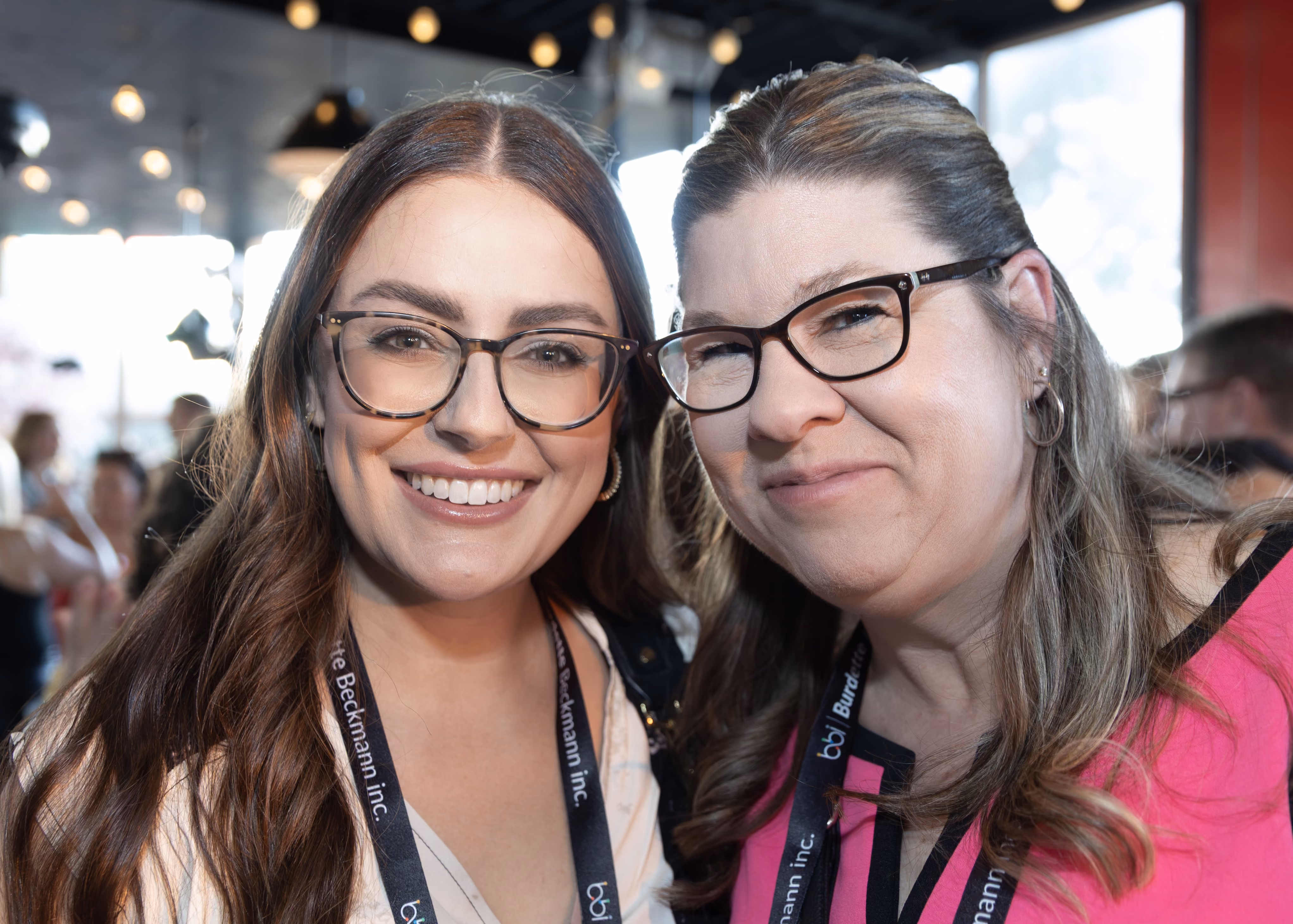 Two women smiling closely together in a well-lit indoor setting, both wearing glasses and conference lanyards, with blurred people and lights in the background.