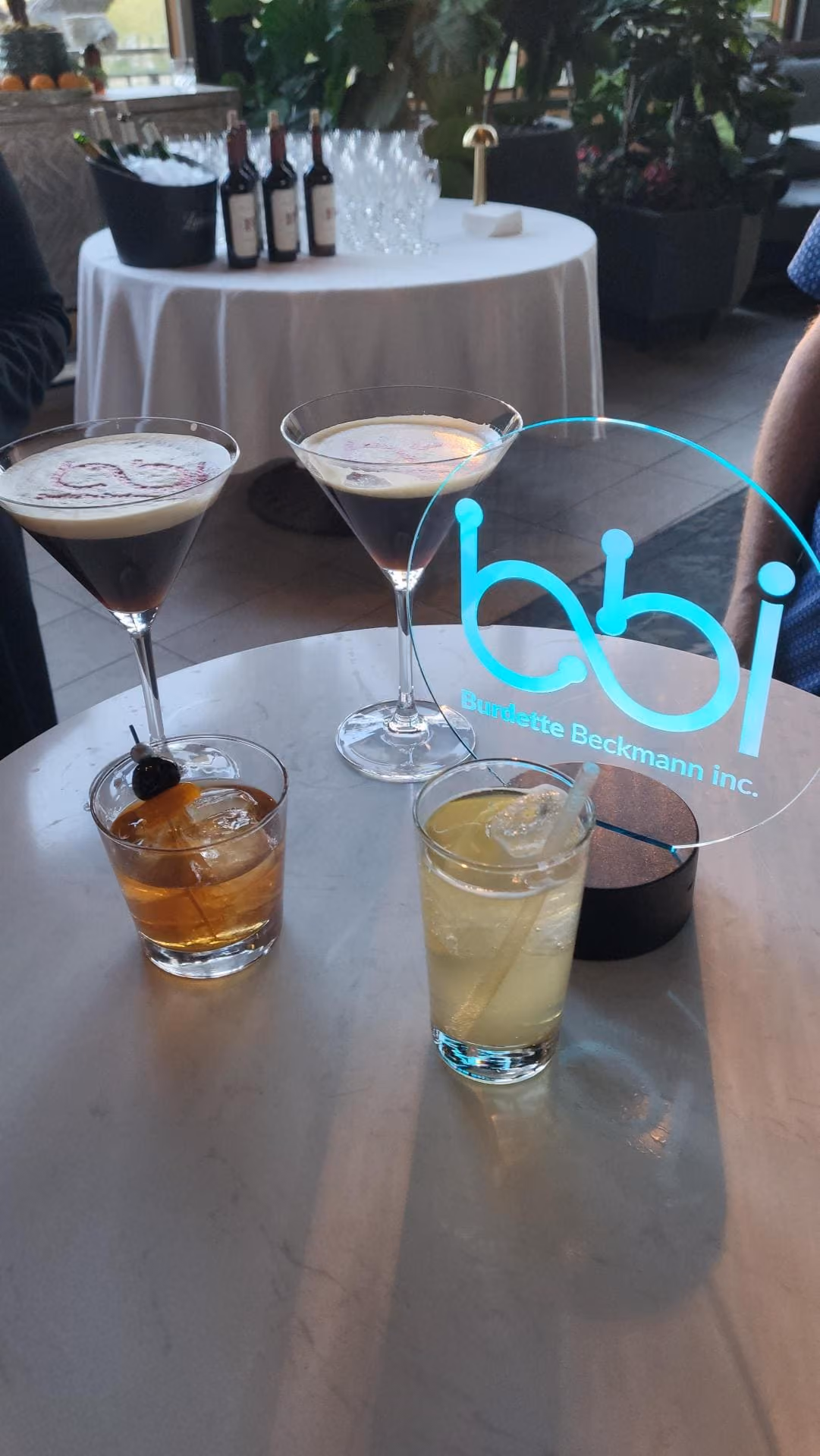 A round marble table with four cocktails: two martinis with foam tops and two highball glasses, one with an orange garnish and the other with a lemon wedge and straw, next to a blue acrylic sign reading 'bbi Burdette Beckmann inc.' In the background, a larger round table with bottles of wine and stacked wine glasses is visible in a plant-filled indoor setting.