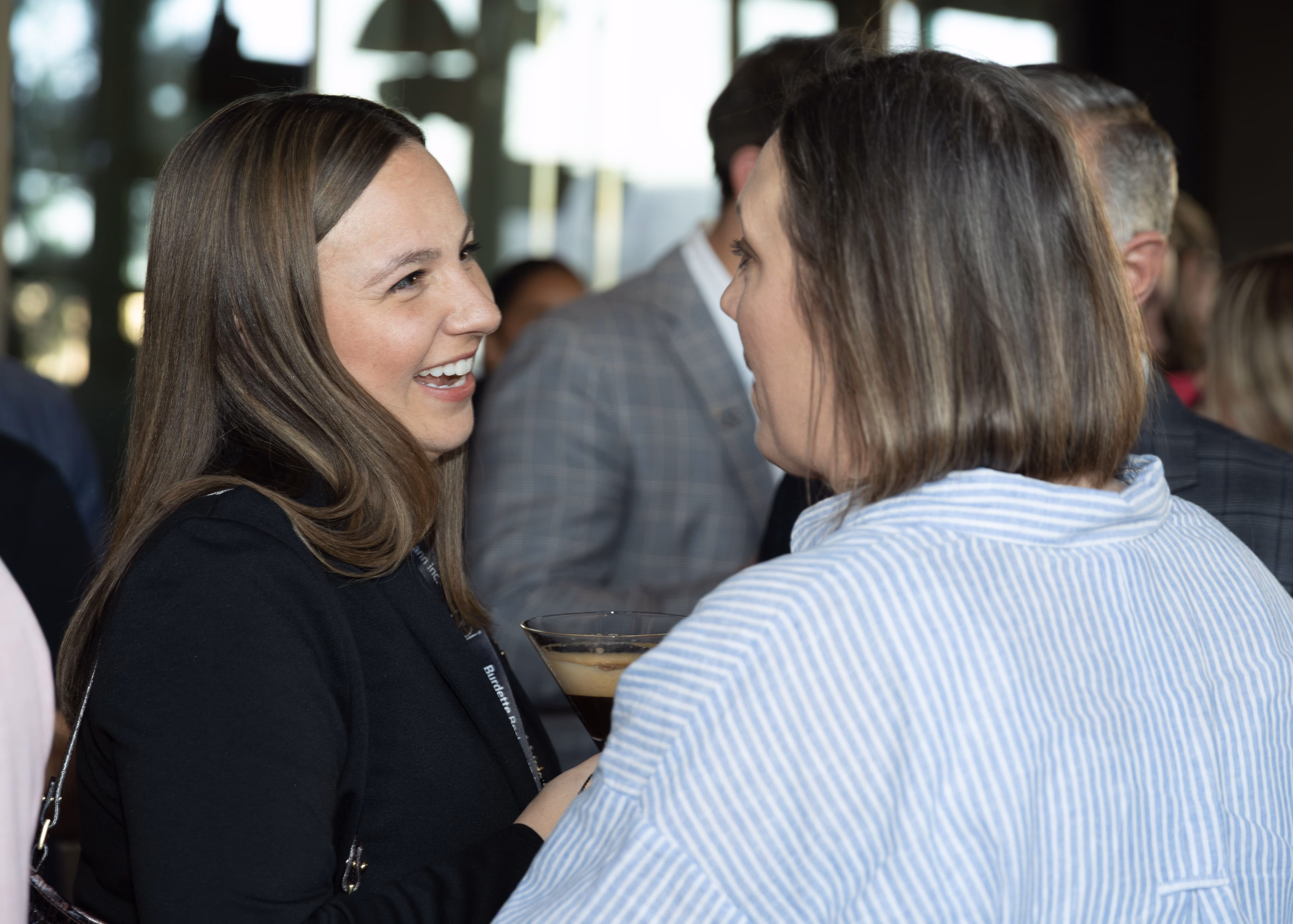 Two women engaged in conversation at a social event, one smiling brightly while holding a drink in a martini glass, background with other people slightly blurred.