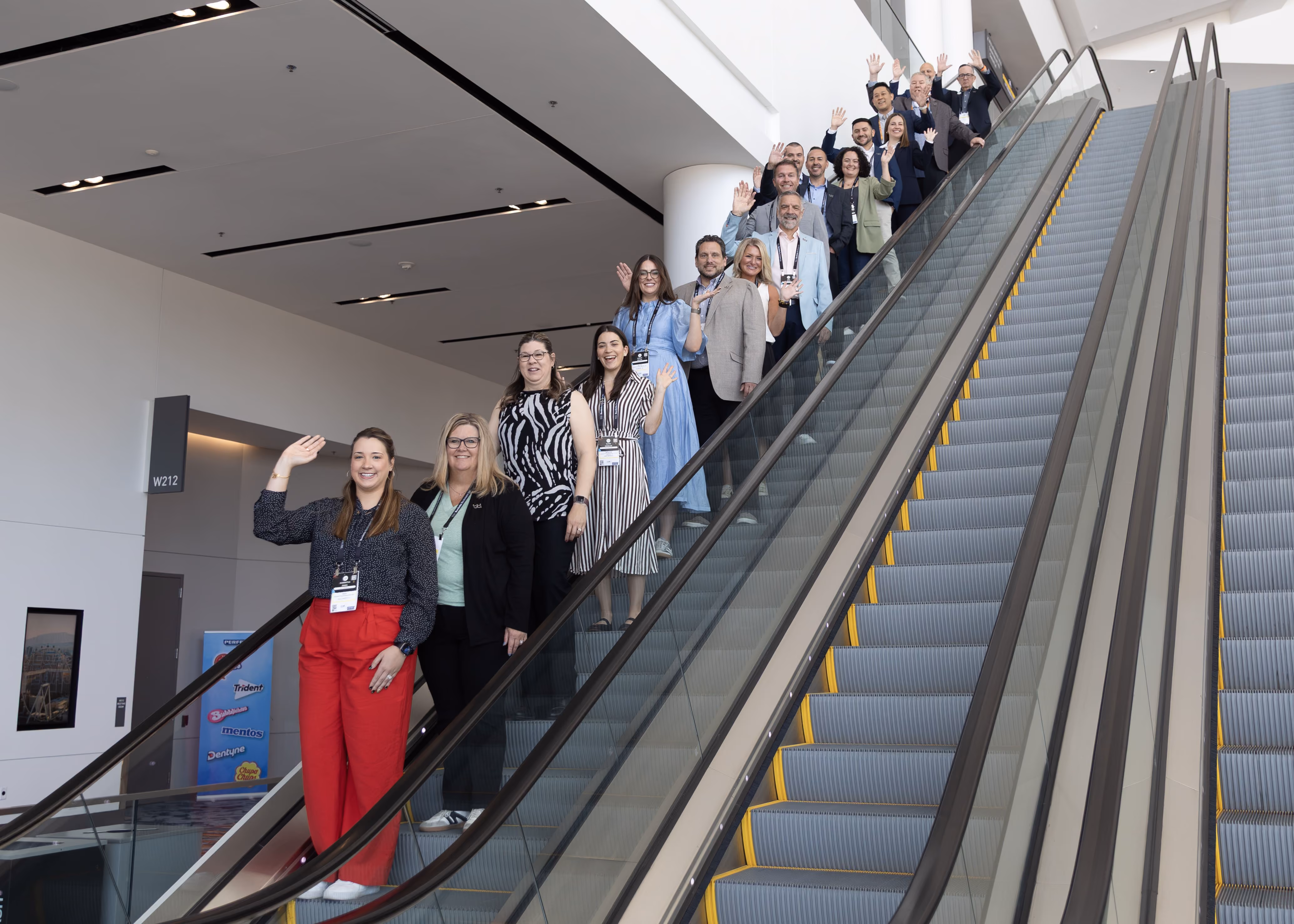 A group of people standing on an escalator indoors, smiling and waving at the camera in a modern, well-lit building.