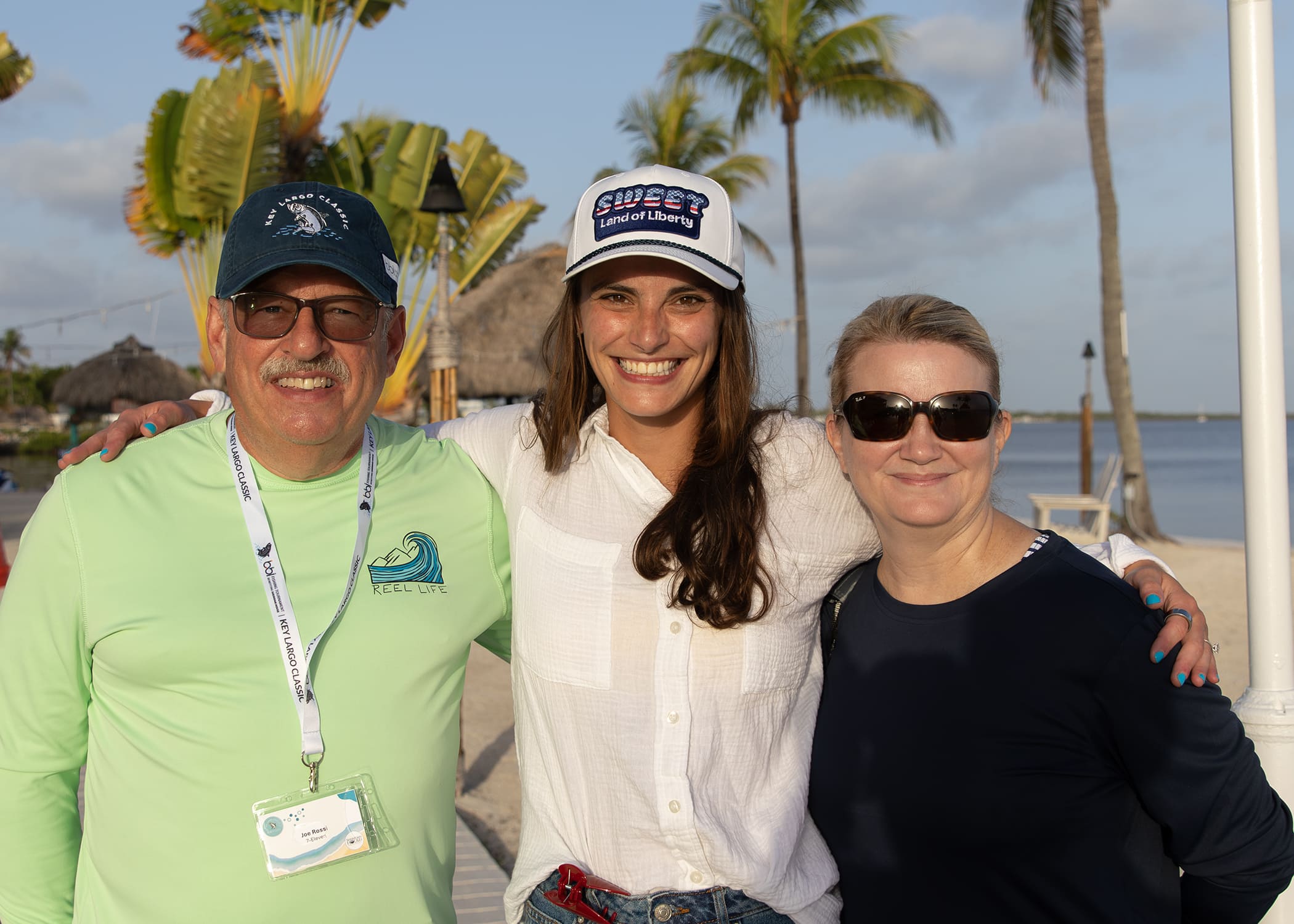 Man in a cap and sunglasses posing with two women outdoors near palm trees.