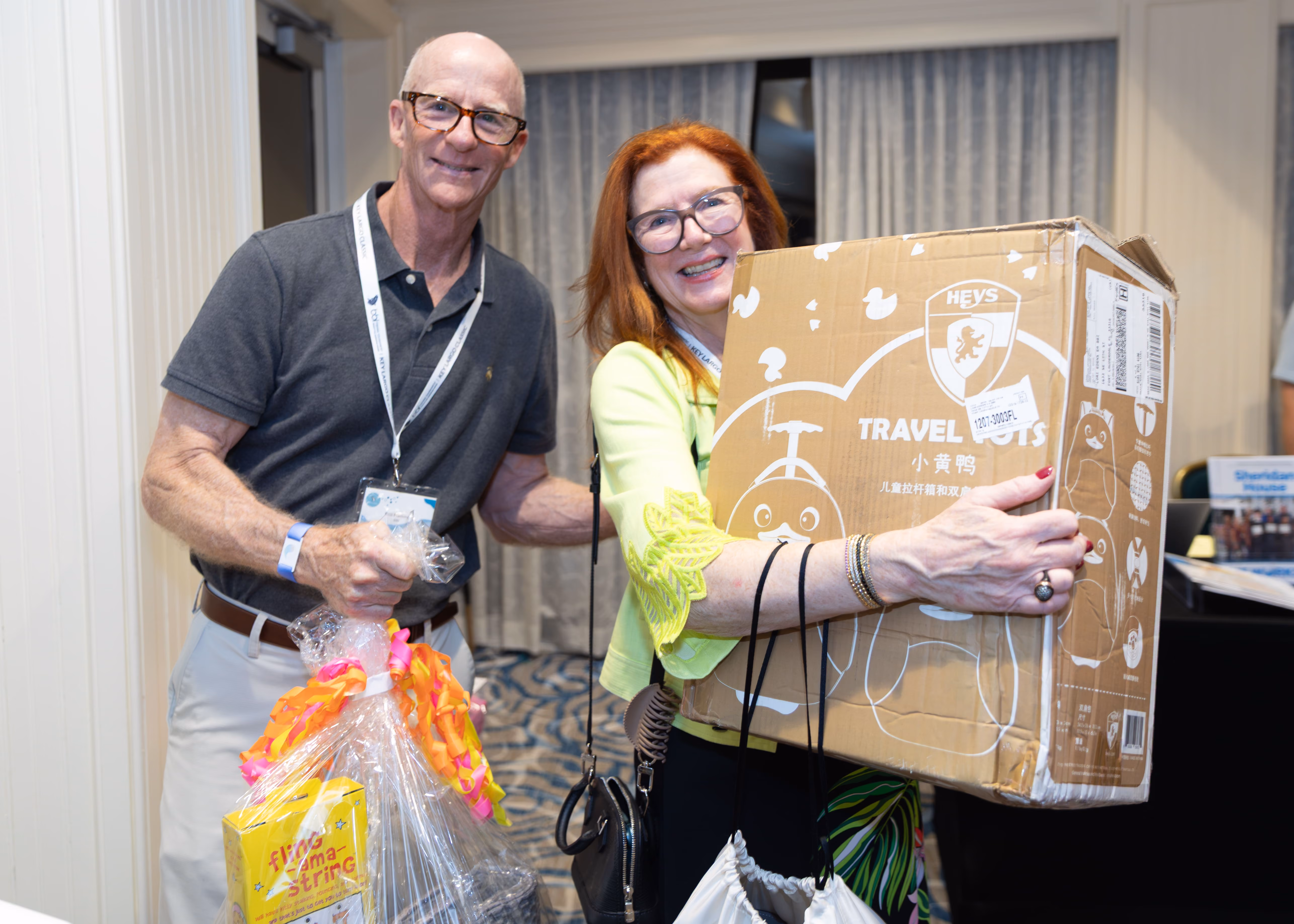 Smiling man and woman holding gift wrapped items, including a large boxed travel toy and packaged gift.