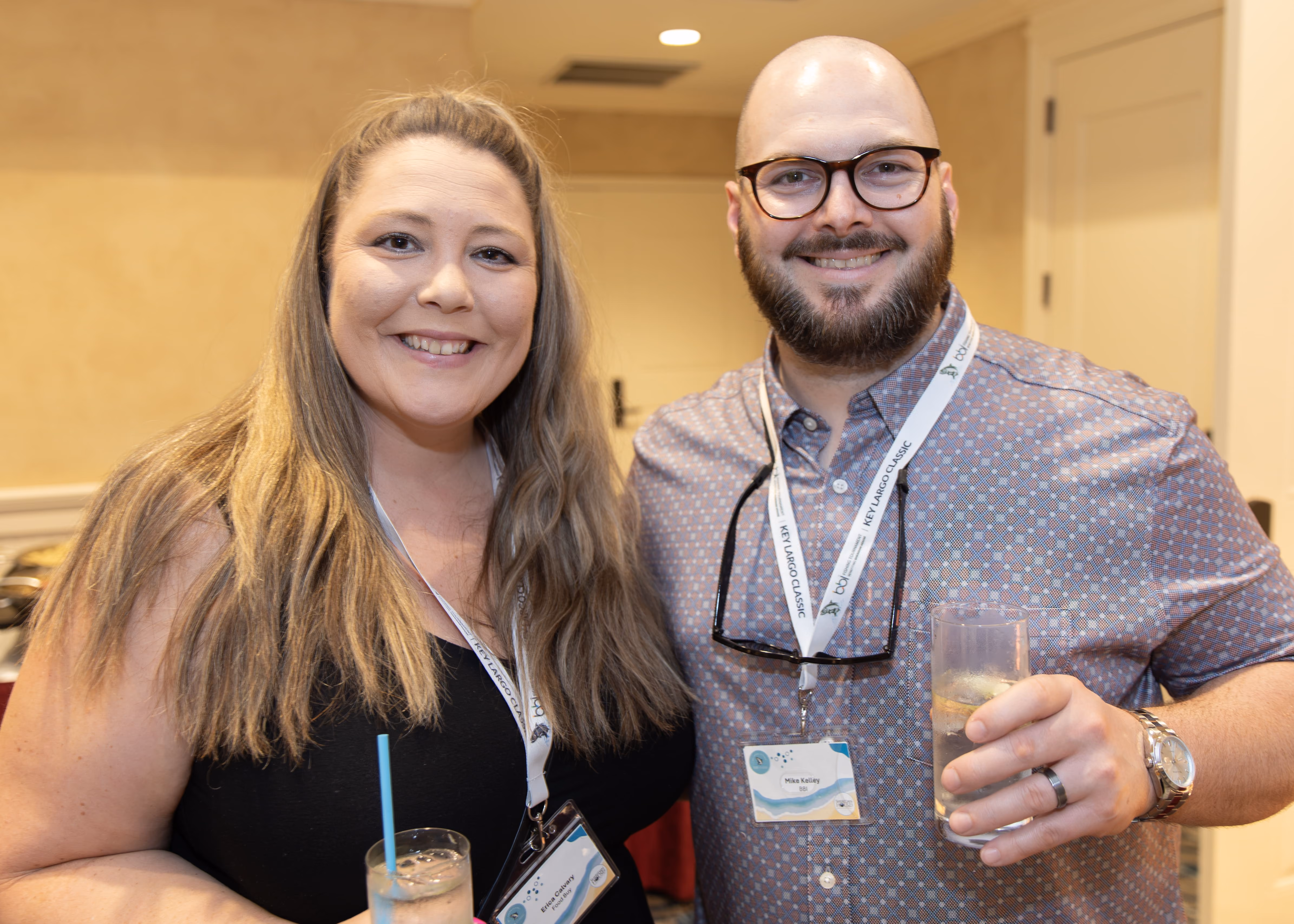 Smiling man and woman wearing conference badges, each holding a drink in a room.