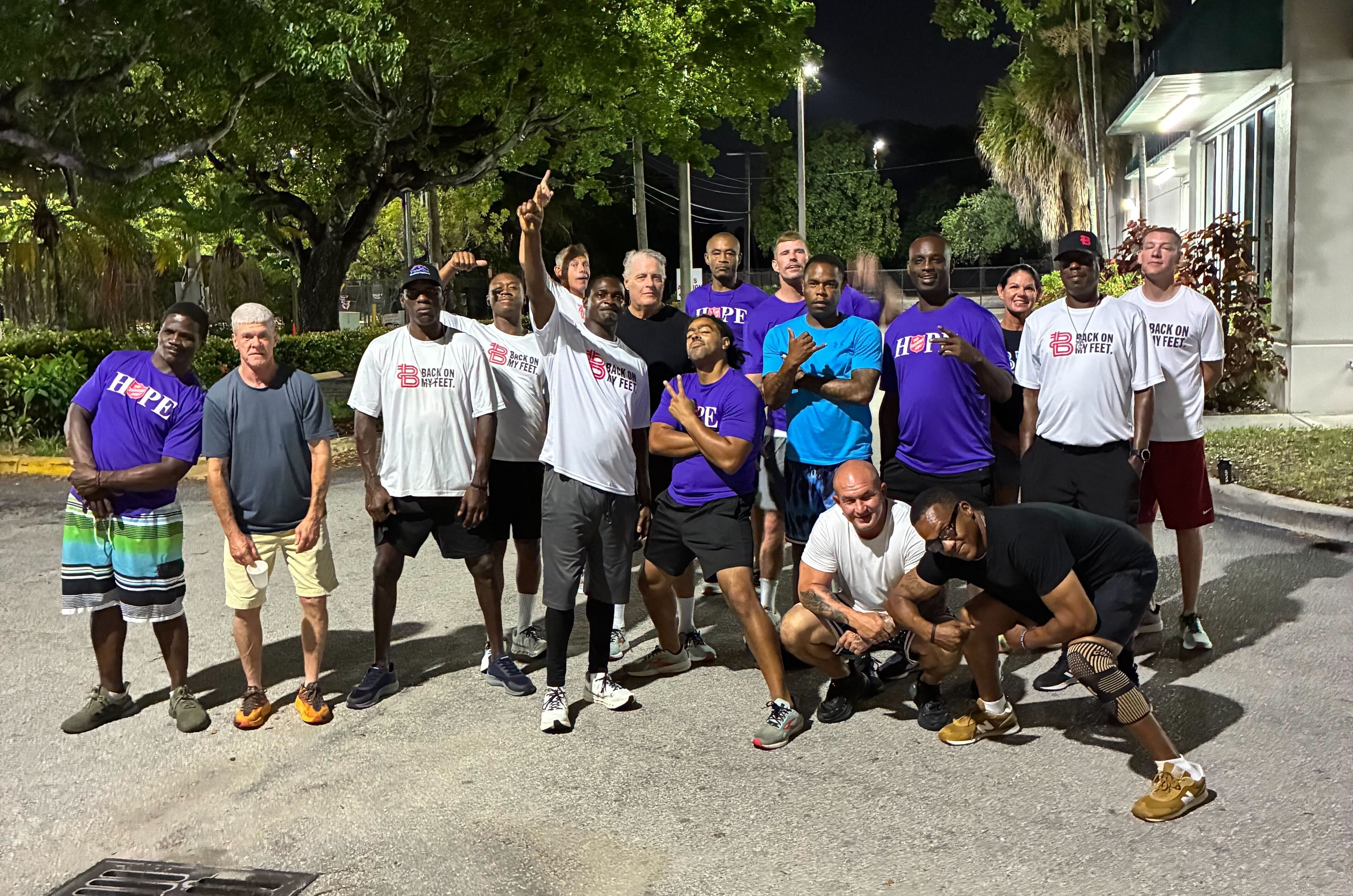 Group of Back on My Feet members in purple shirts posing together outdoors at night, one raising a fist