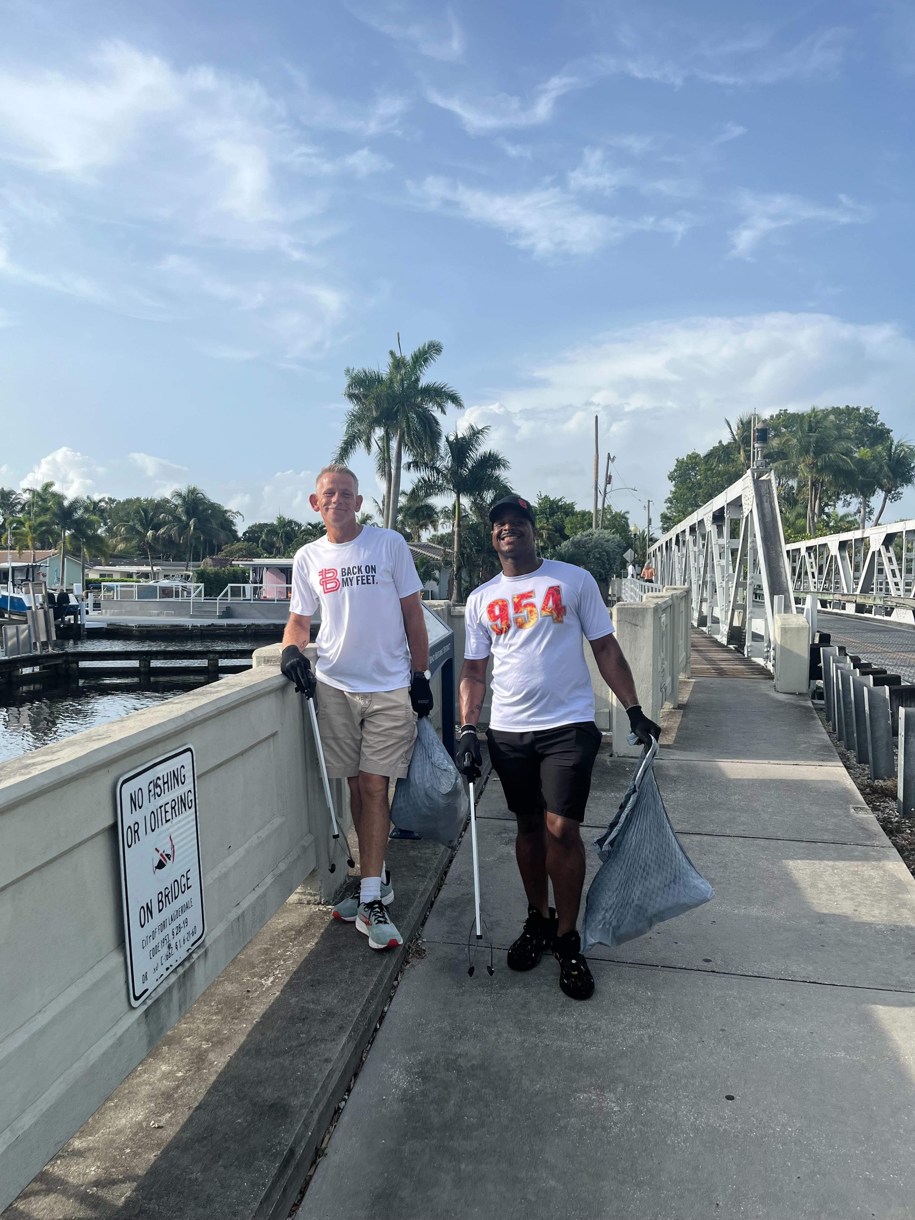 Two men carrying trash bags while walking along a waterfront wall during a community clean up