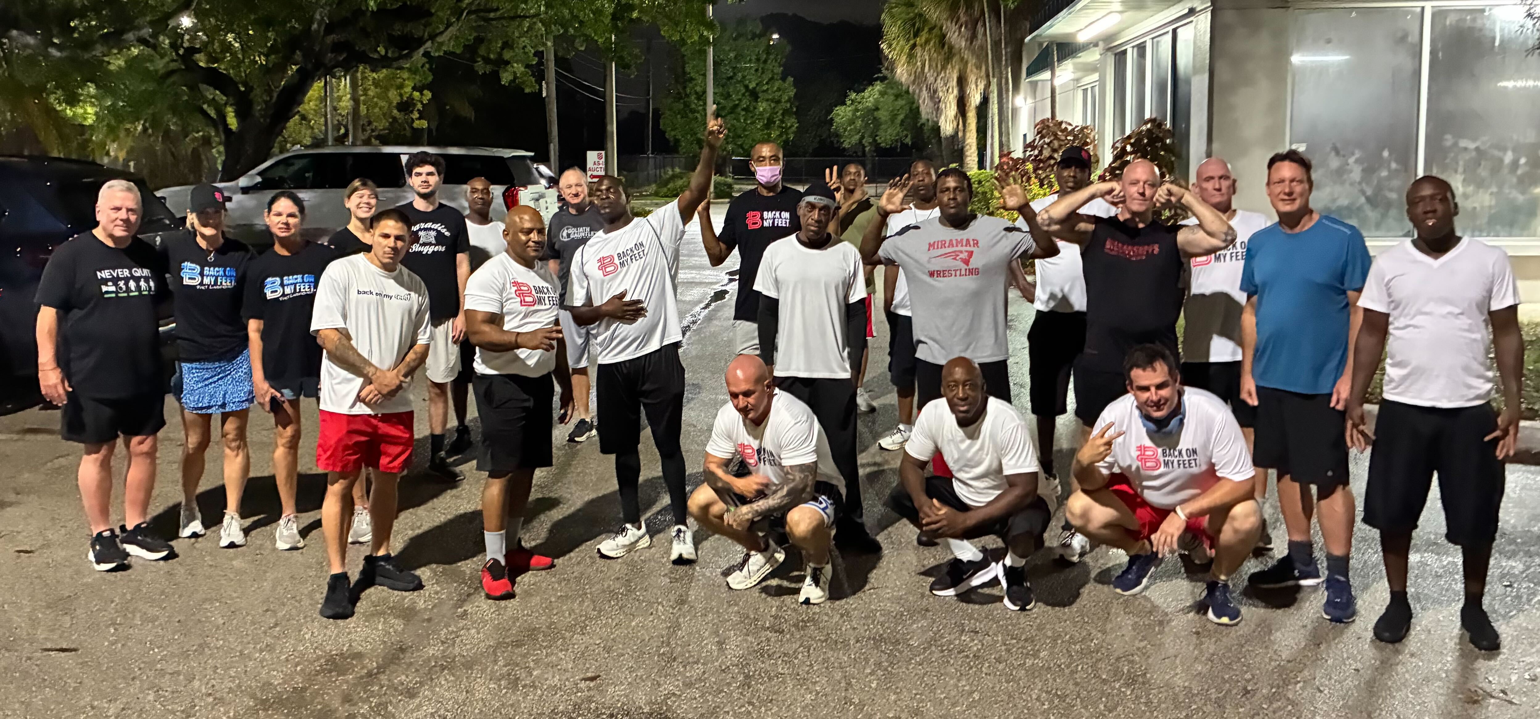 Group of Back on My Feet members in white shirts posing together outdoors at night, some raising their arms