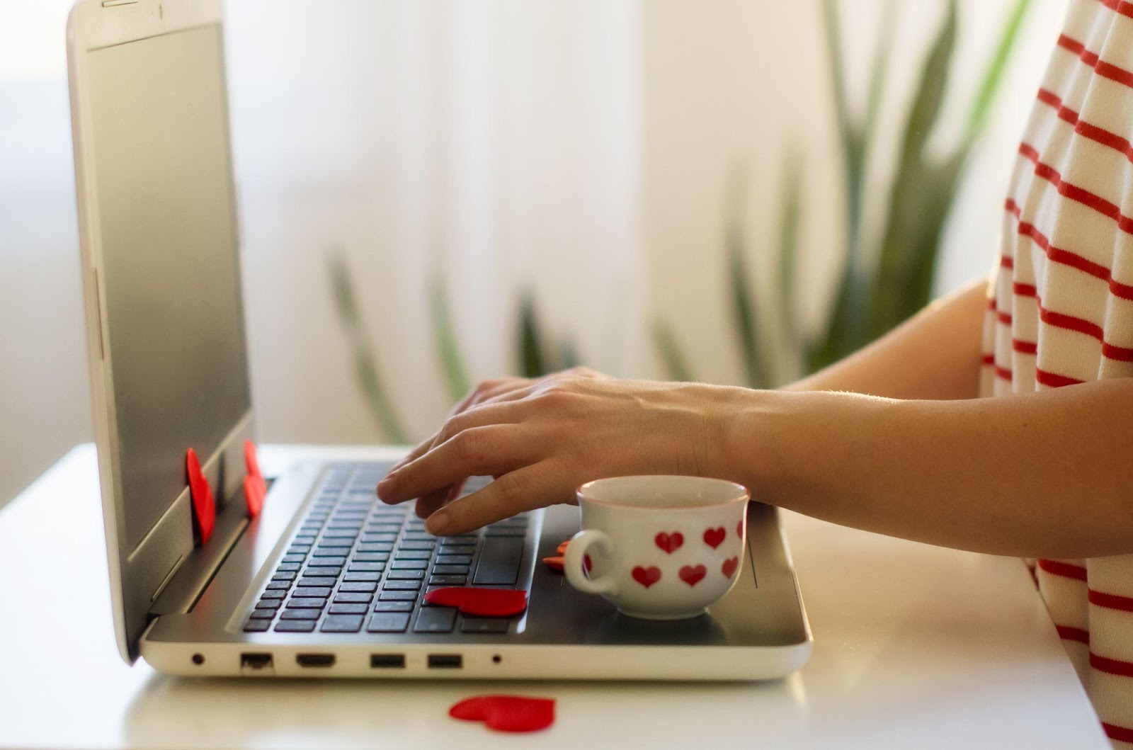 A cup with heart designs is next to a person using a laptop covered in heart confetti.