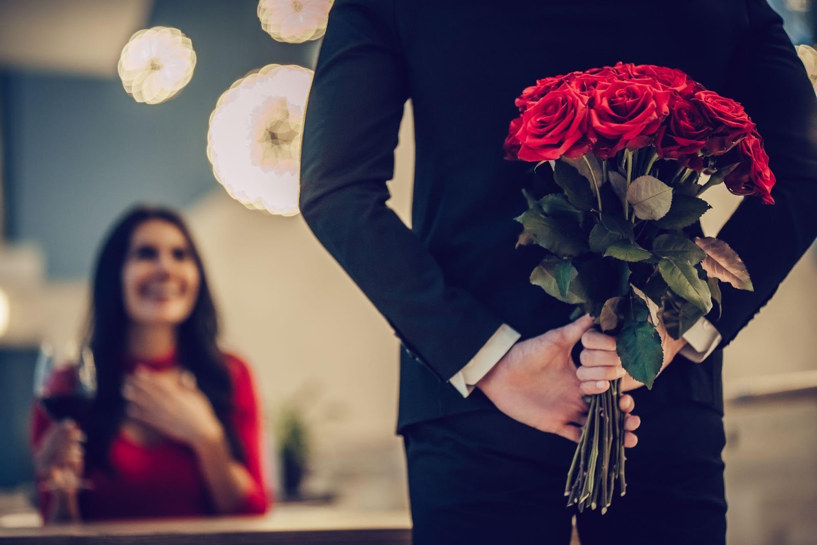A man hiding roses behind his back surprises his Valentine’s Day date.