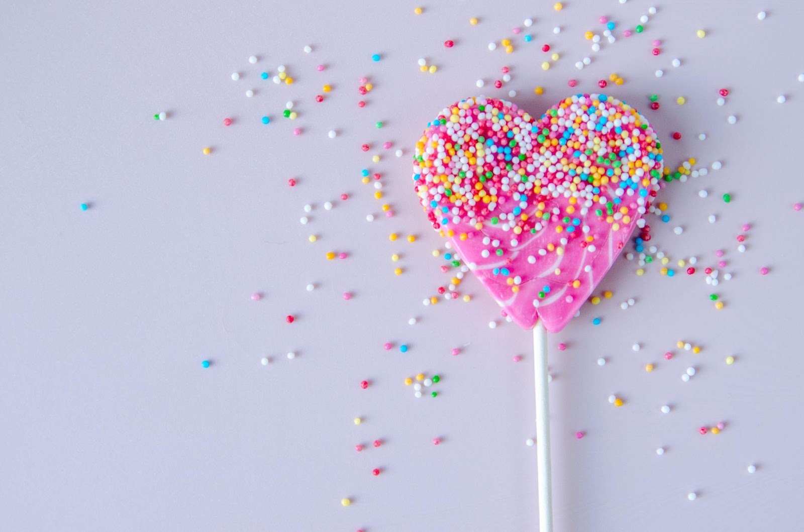 Colorful candy confetti sprinkled on a pink, heart-shaped lollipop.