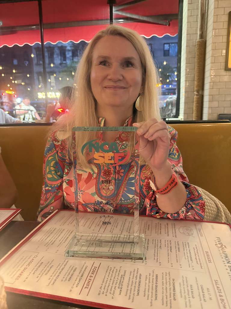 Smiling woman in a restaurant booth holds a glass NCASEF Broker Of The Year award engraved BBI 2026.
