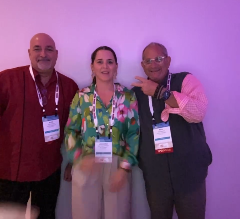 Two men and a woman wearing lanyards pose against a pink wall, the man at right in a bbi vest.