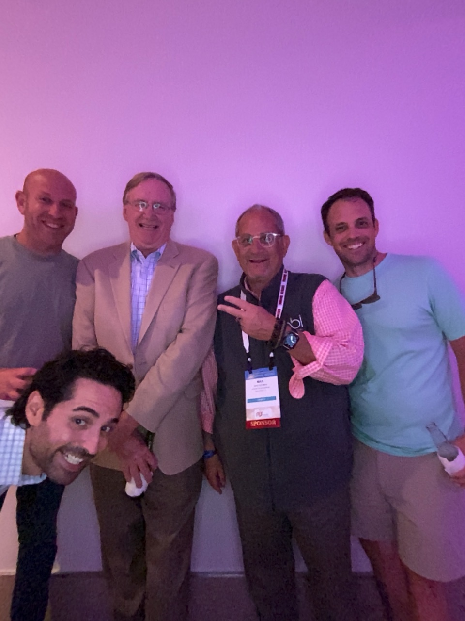Five men pose against a pink wall, one in a vest with a bbi logo and a Sponsor badge, flashing a peace sign.