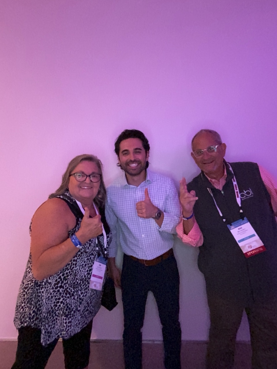 A woman and two men give thumbs up against a pink wall, the man at right in a bbi vest with a Sponsor badge.