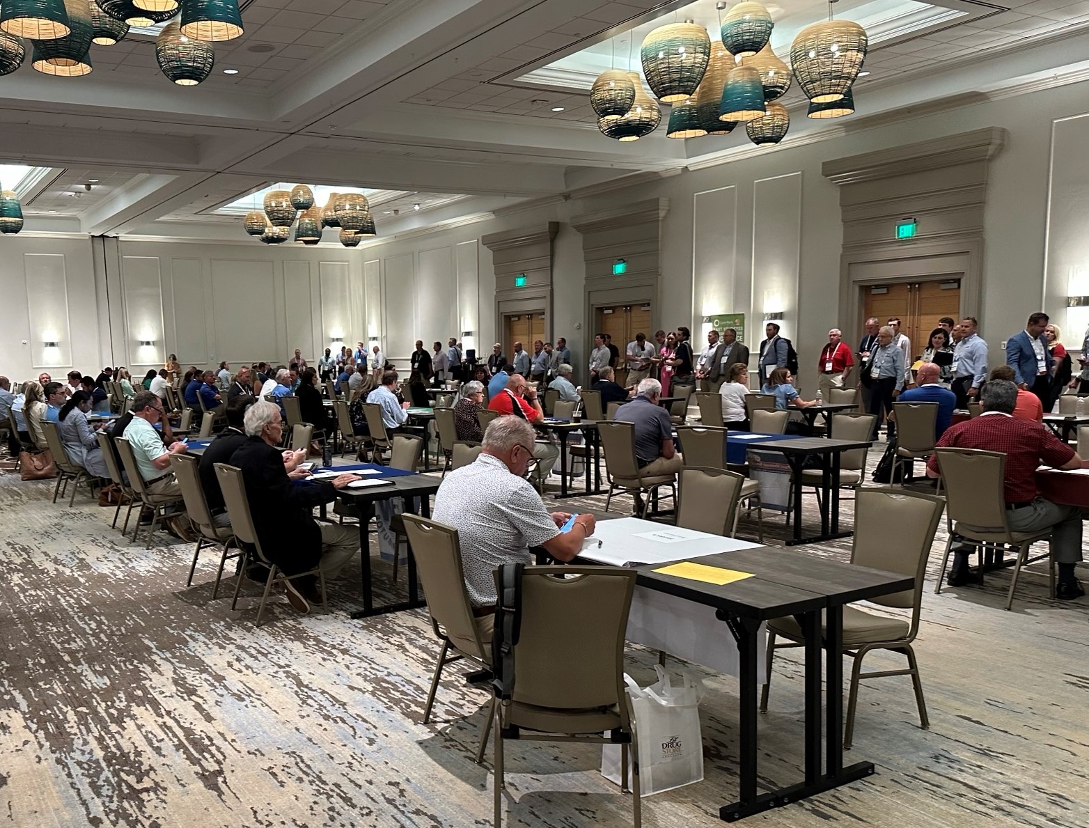 Attendees seated at small tables in a hotel ballroom with woven pendant lights, others standing at the back.