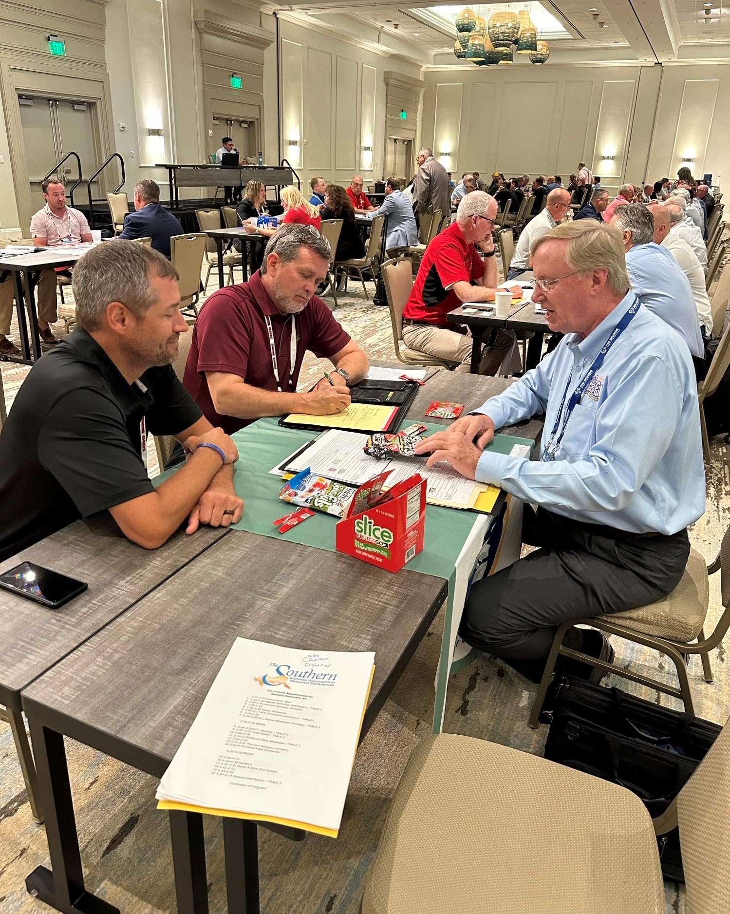 Three men talk across a table with papers and a red Slice box, with other one-on-one meetings behind them.