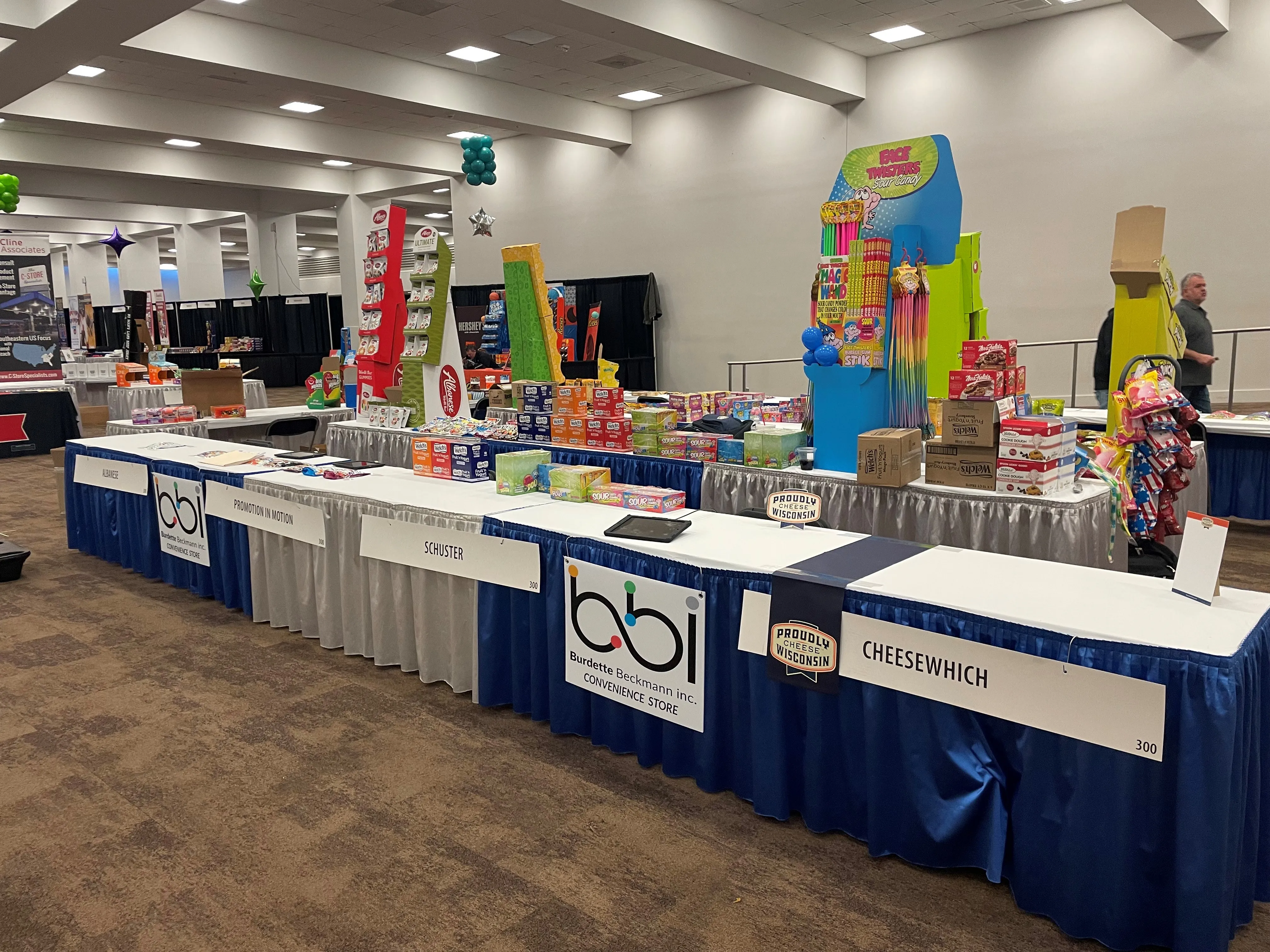 Vendor tables with a bbi Convenience Store sign beside Schuster and Cheesewhich placards and candy displays.
