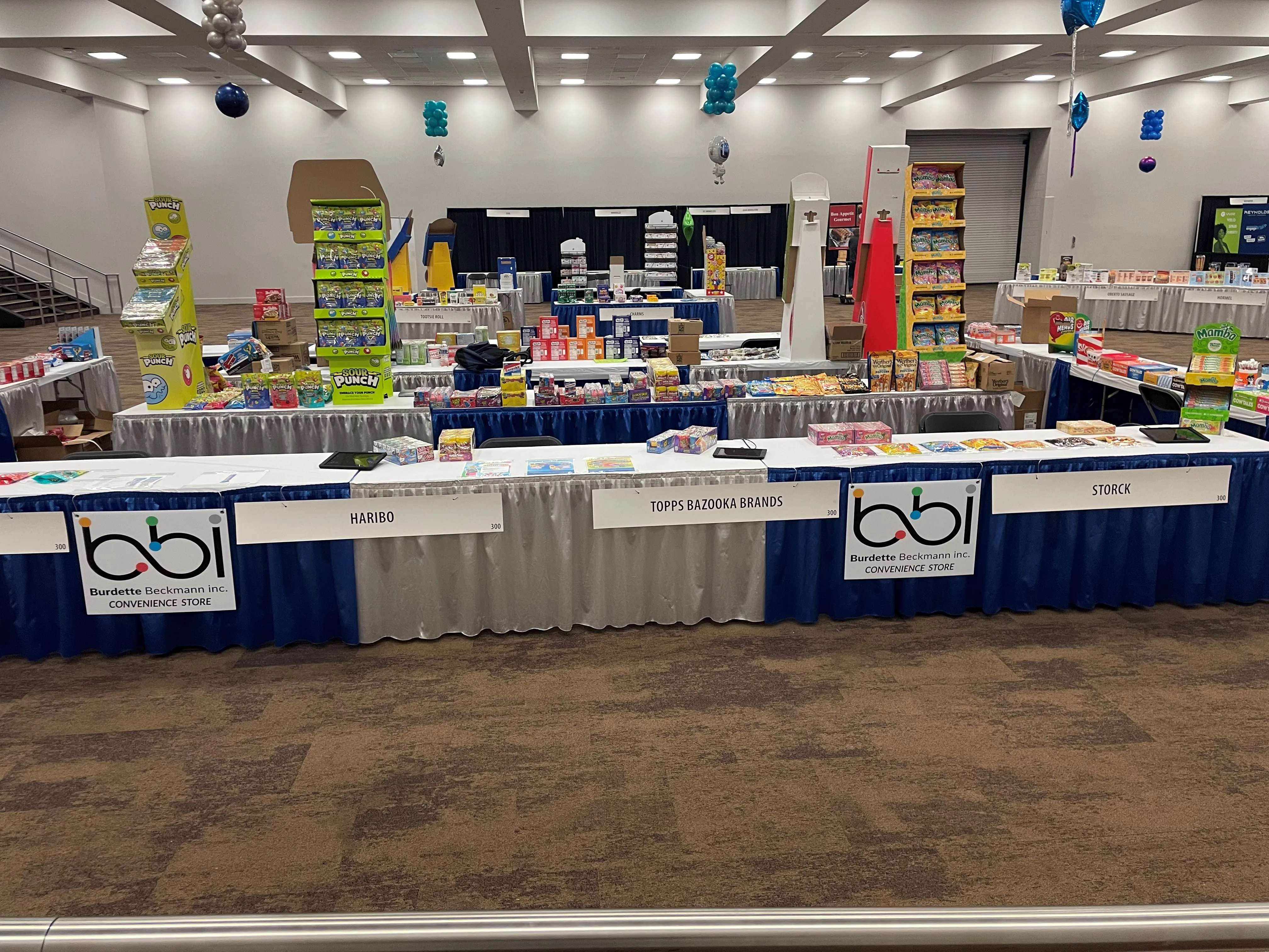 Vendor tables with bbi Convenience Store signs and Haribo, Topps Bazooka Brands and Storck placards in a large hall.