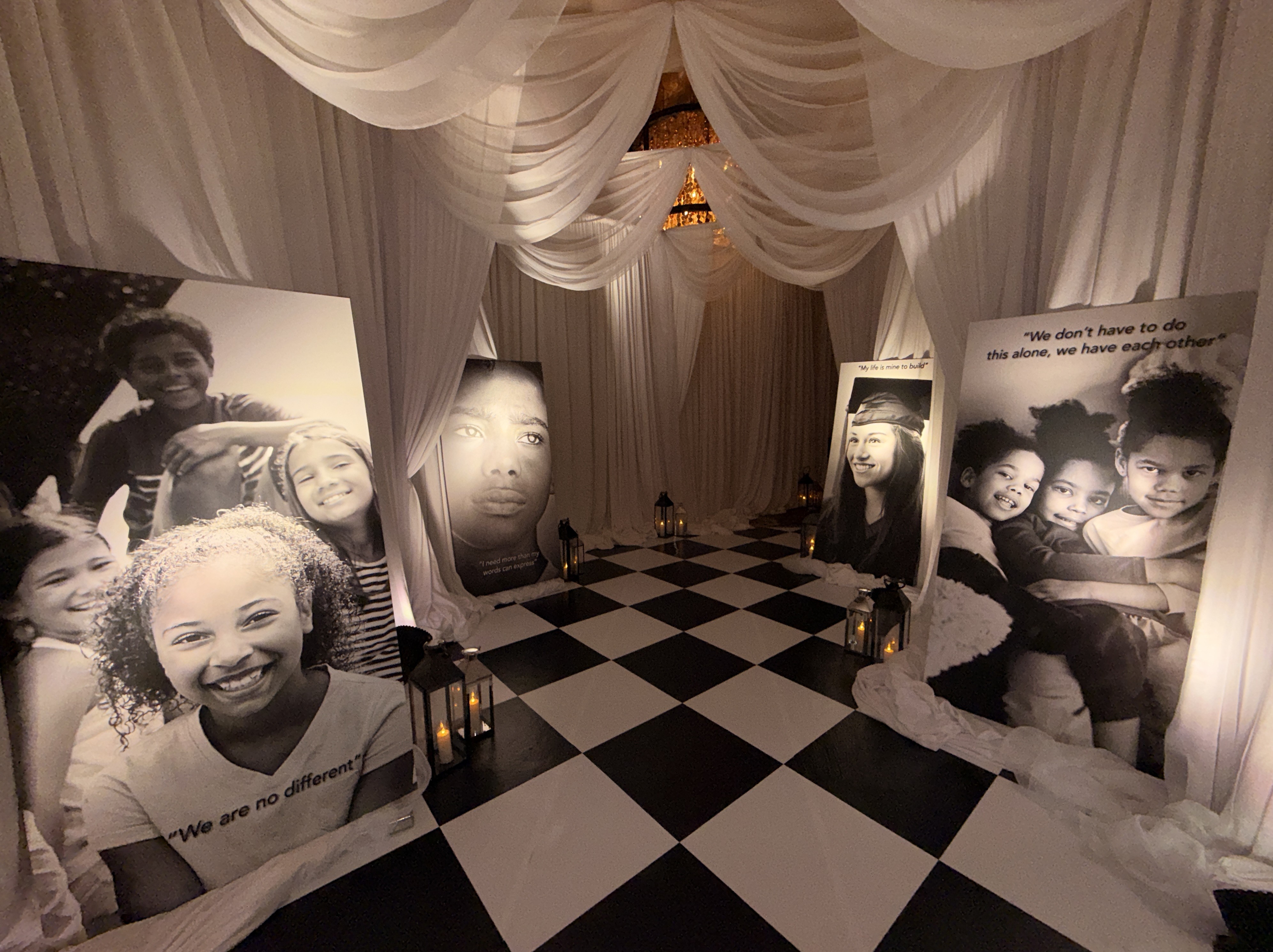Large black-and-white portraits of smiling children and lanterns on a checkerboard floor under draped white fabric.
