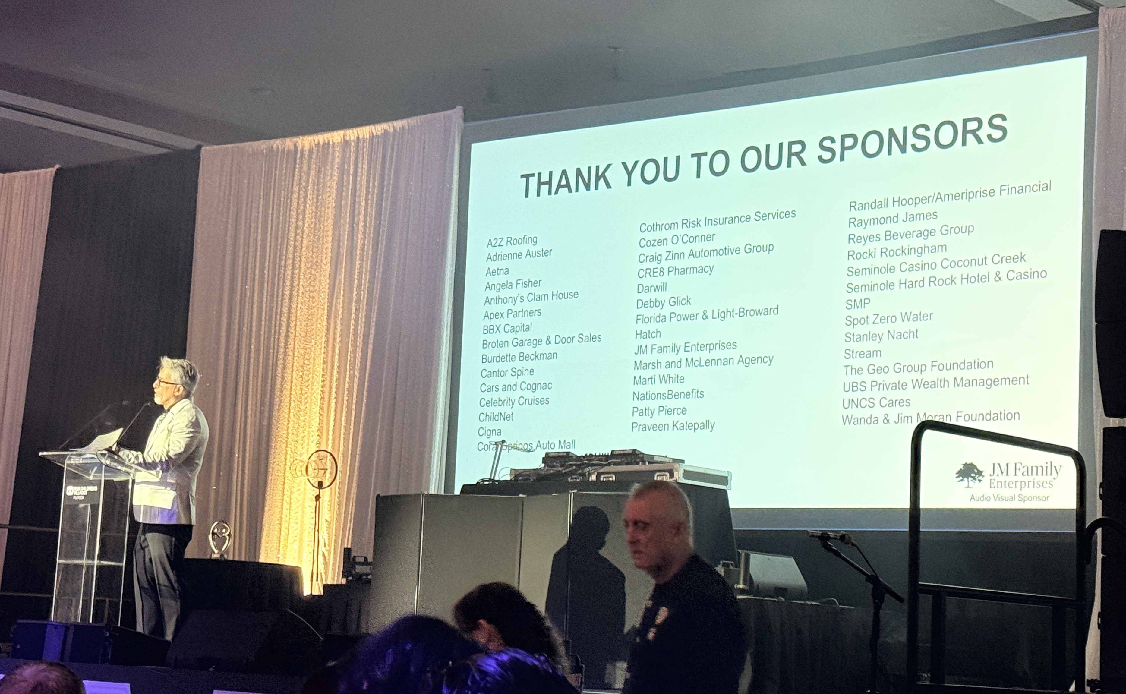 Man at a podium beside a projected slide reading Thank You to Our Sponsors, with columns of sponsor names.