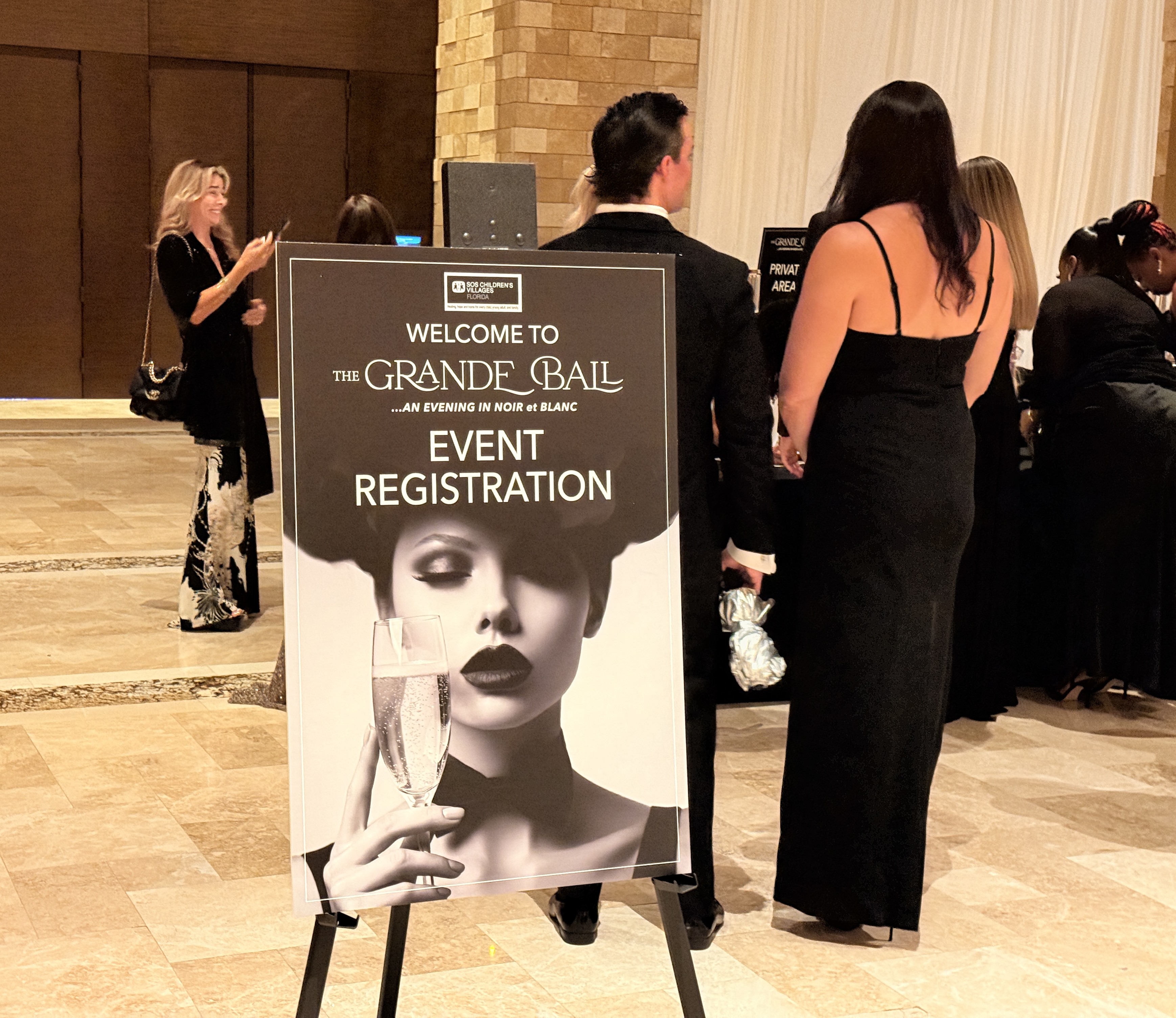Easel sign reading Welcome to The Grande Ball, Event Registration, with guests in evening wear standing behind it.