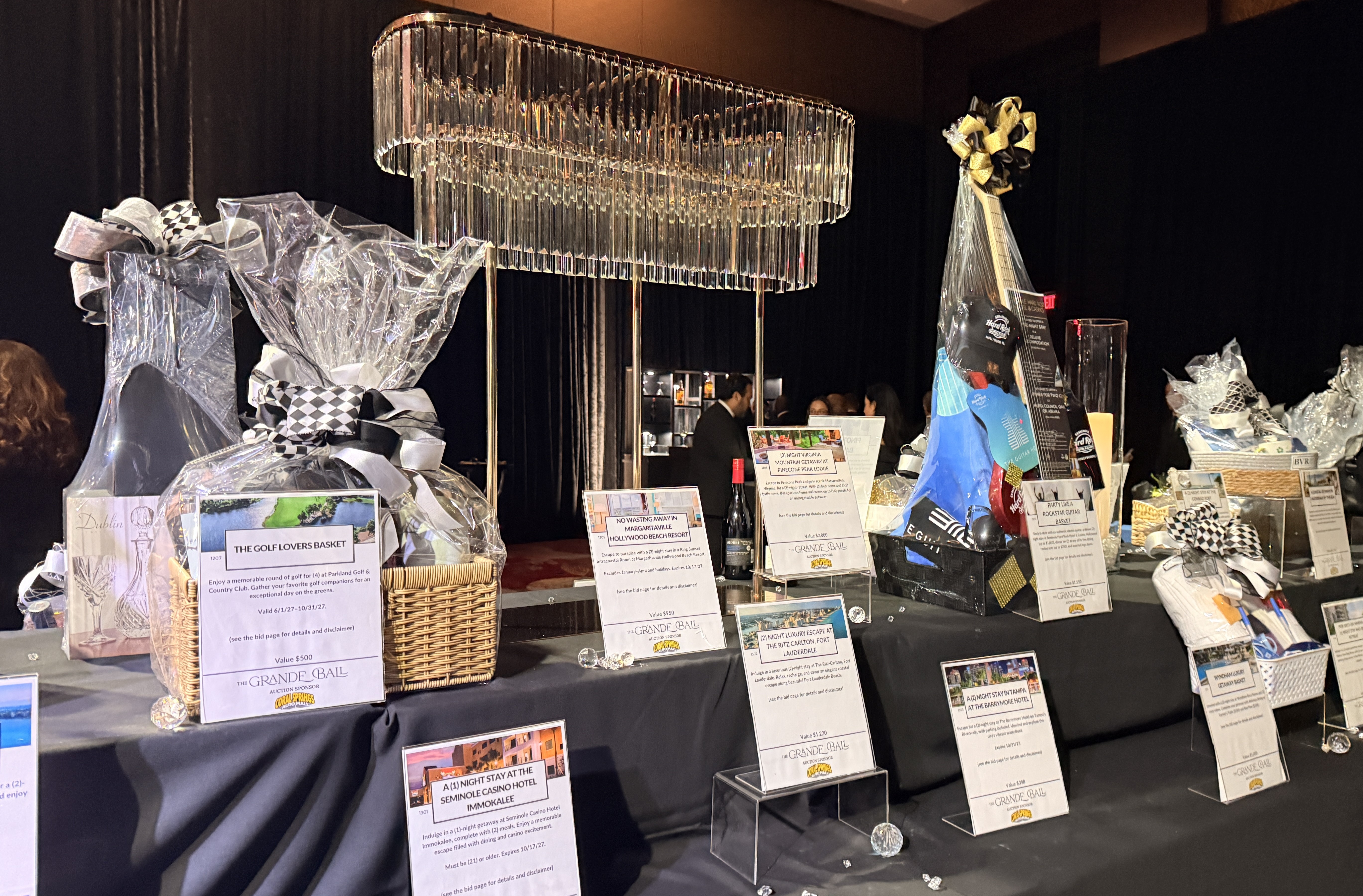 Auction gift baskets with printed description cards on a black-draped table under a crystal chandelier.
