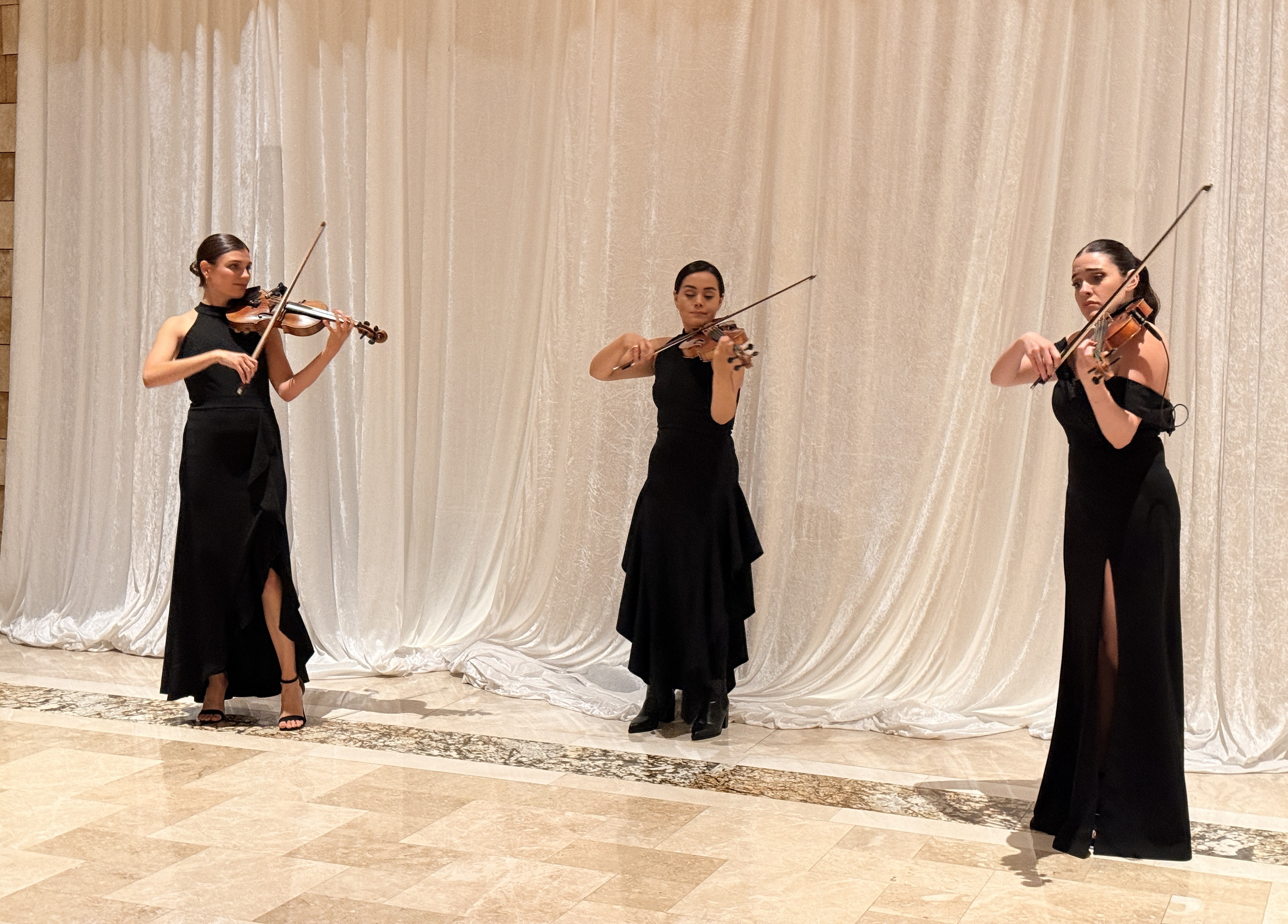 Three women in black gowns playing violins in front of a white draped curtain.