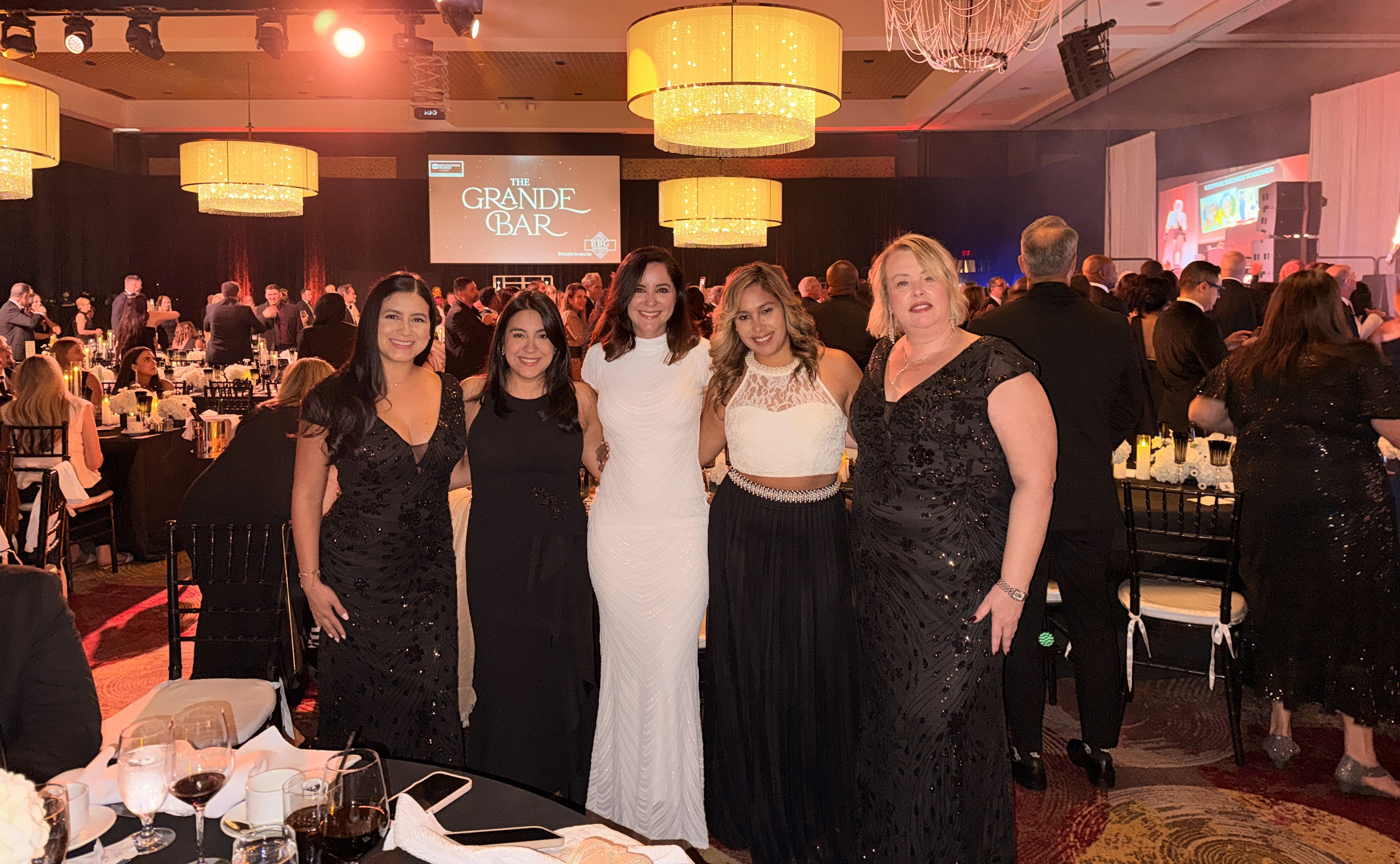 Five women in black and white gowns stand together at a dining table in a crowded ballroom.
