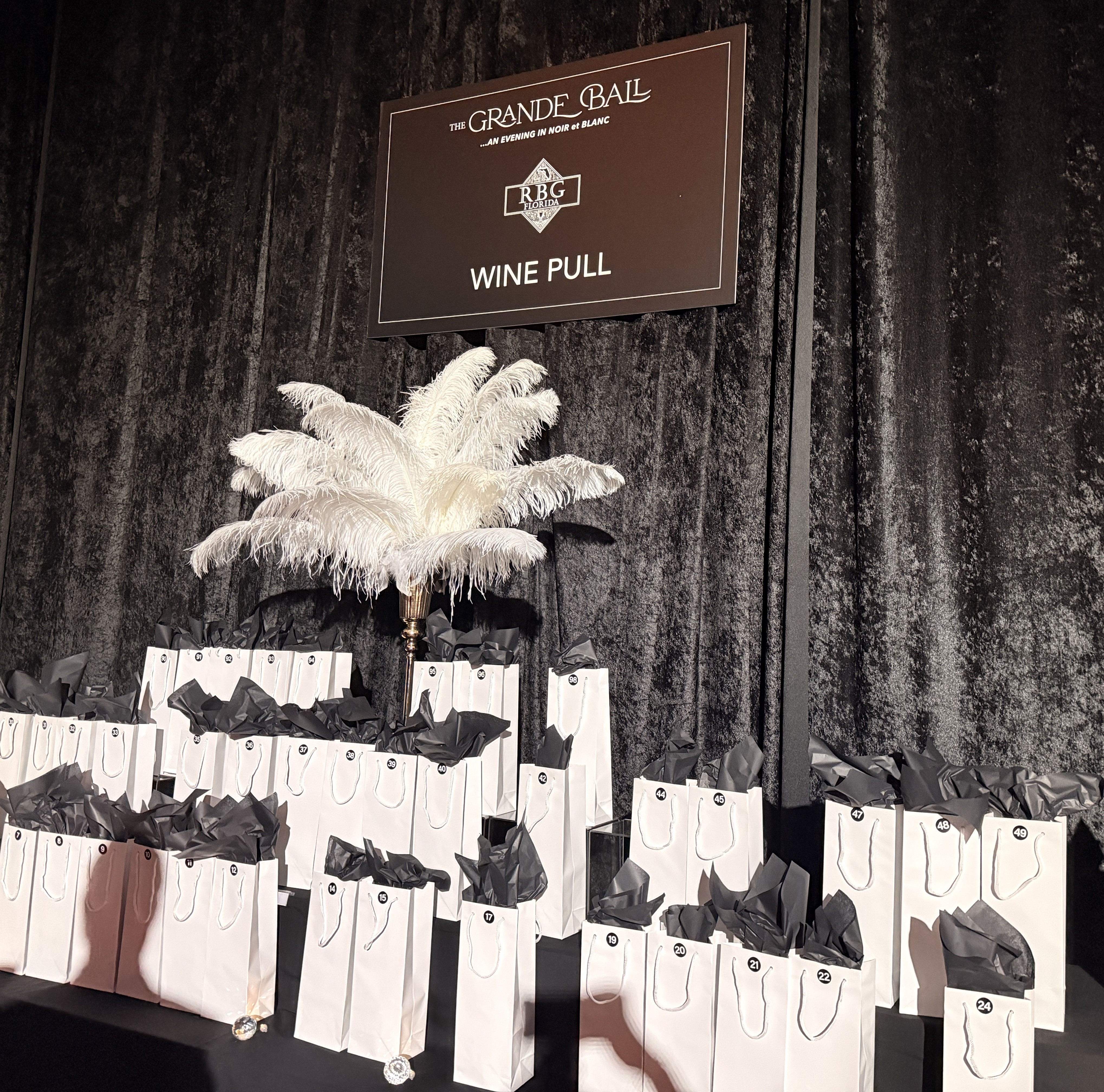 Rows of white numbered gift bags with black tissue under a Wine Pull sign and a white feather centerpiece.