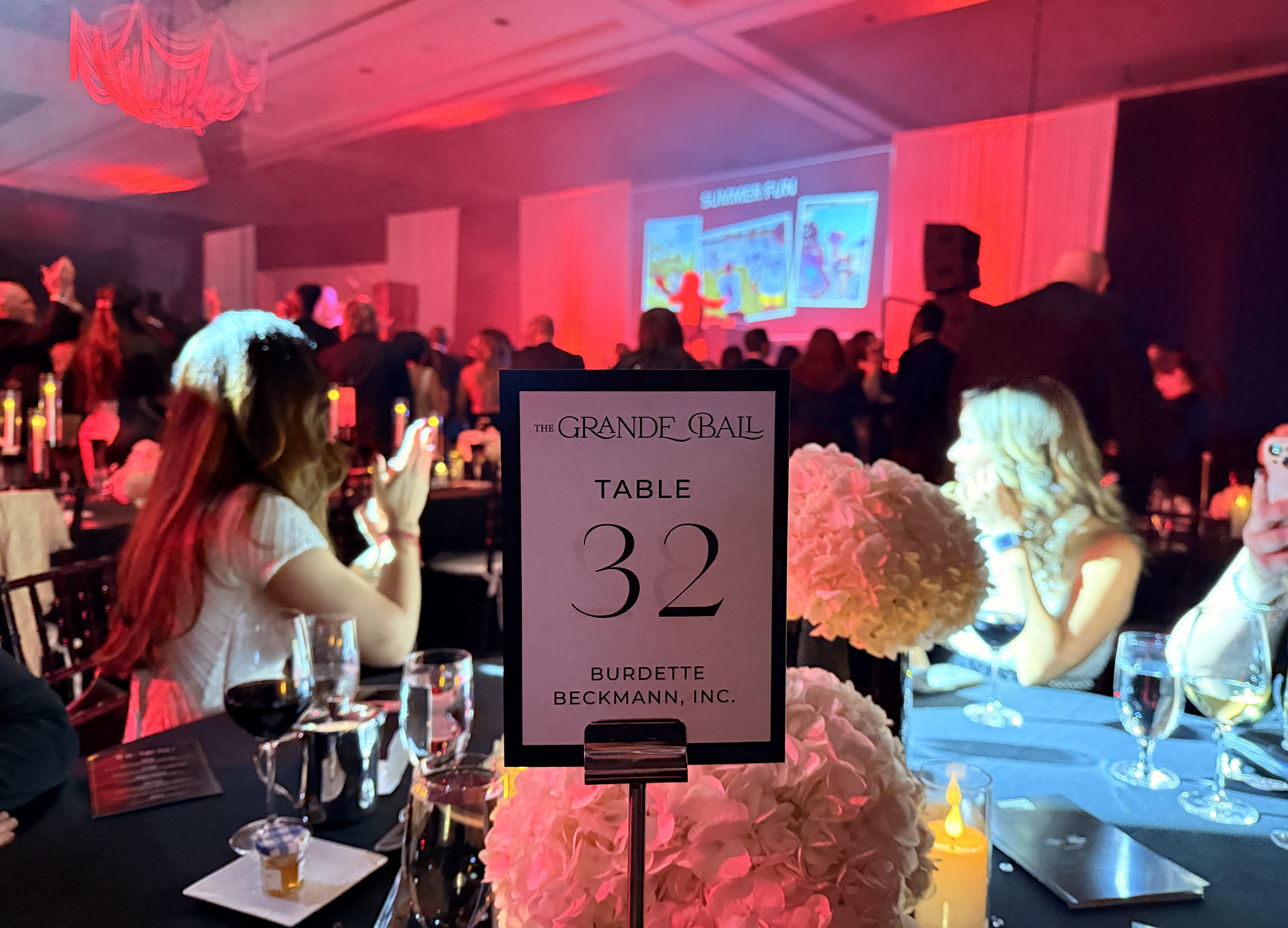 Table sign reading Table 32 Burdette Beckmann, Inc. in a red-lit ballroom with guests facing a projection screen.