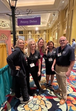 Five people in black shirts and name badges pose beneath a purple Welcome Petrey Drive sign in a carpeted hall.