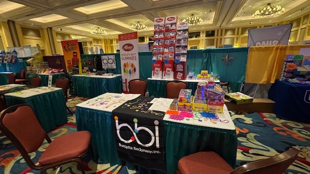 bbi booth table with candy boxes and bead necklaces in front of an Albanese World's Best Gummies banner and empty chairs.