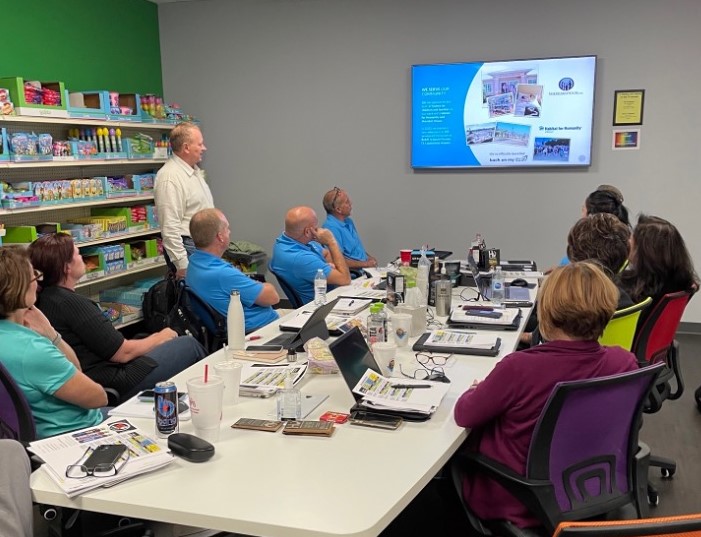 Attendees at a conference table watch a slide about Habitat for Humanity as a man stands beside shelves of candy displays.