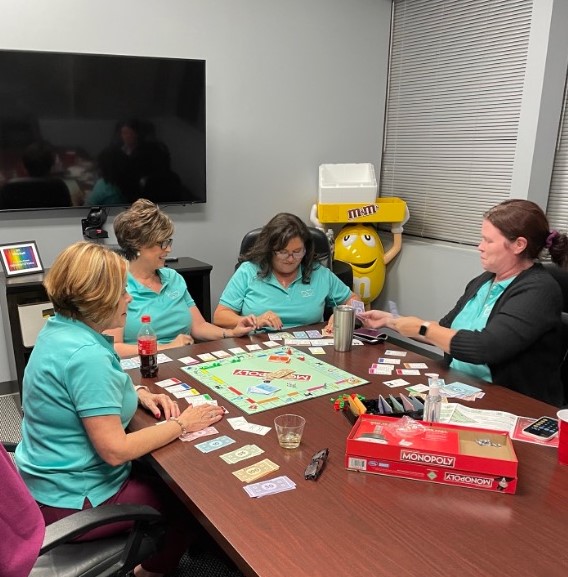 Four women in teal polo shirts play Monopoly at a conference table with an M&M's figure behind them.