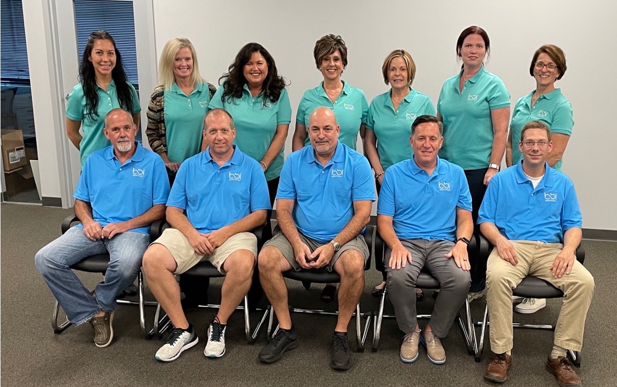 Twelve BBI staff in blue and teal polo shirts pose in two rows, five men seated in front and seven women standing behind.