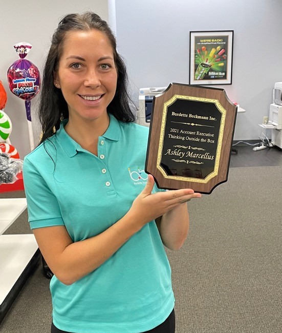 Smiling woman in a teal BBI polo holds a plaque reading 2021 Account Executive Thinking Outside the Box, Ashley Marcellus.