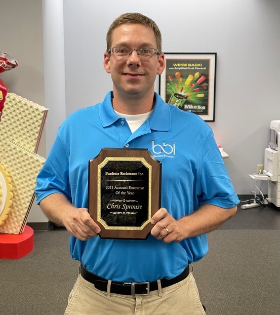 Man in a blue BBI polo holds a plaque reading 2021 Account Executive Of the Year, Chris Sprouse.