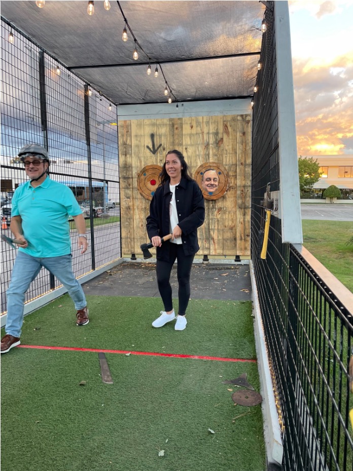 Woman holds an axe in an outdoor throwing lane with wooden targets as a man in a teal shirt and helmet walks past.