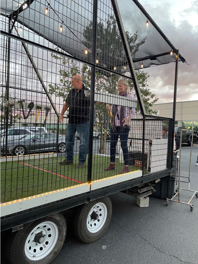 Man and woman stand on turf inside a caged axe-throwing trailer, seen from outside, with axes hanging on a rack.