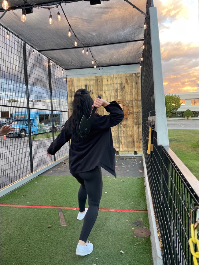 Seen from behind, a woman in black winds up to throw an axe at a wooden target in a caged lane at dusk.