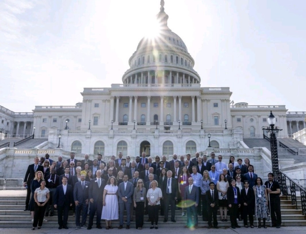 Large group gathered on the steps of the U.S. Capitol, with the dome and bright sun above them.